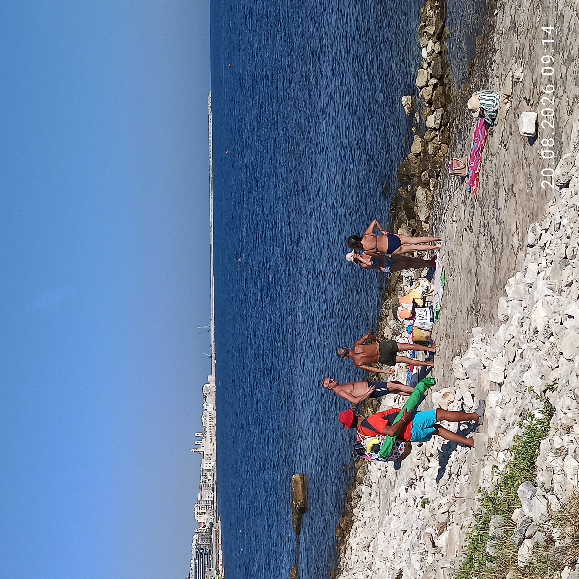 The image depicts a rocky beach with several people enjoying the water and the coastline. In the foreground, a person wearing a red shirt and blue shorts walks with beach gear. Further back, a group of four individuals, two men and two women, are gathered near the water's edge. The women are wearing bikinis, and one of the men is shirtless. Behind them, various beach accessories like bags and towels are laid out. The background reveals the city of Molfetta, Italy, with its characteristic buildings and a prominent lighthouse. The sea is calm, and the sky is clear and blue. The overall scene conveys a relaxed summer day at the beach.