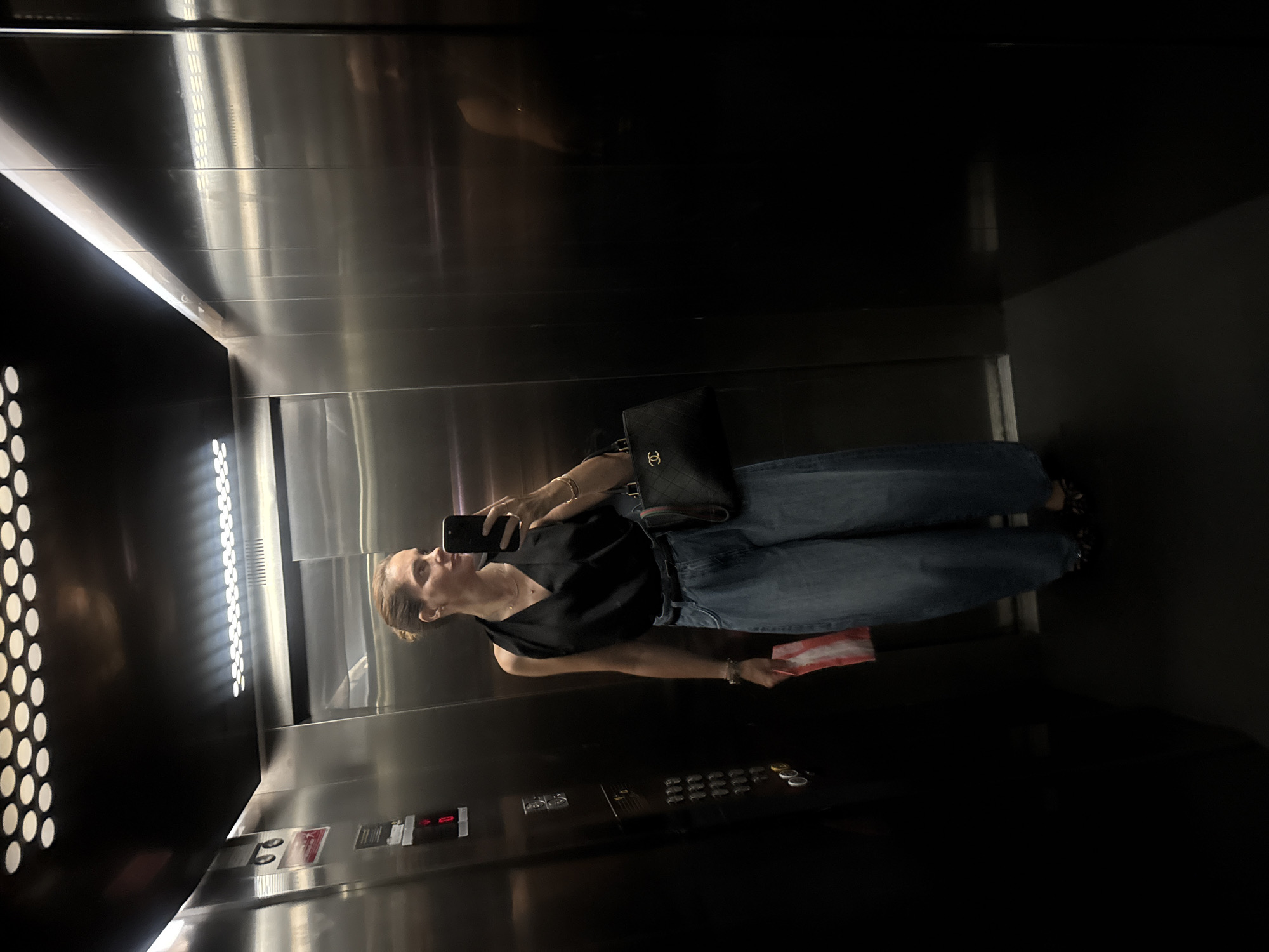 A woman is standing in an elevator, taking a selfie with her phone. She is wearing a black sleeveless top, blue wide-leg jeans, and black patterned shoes. She is holding a red and white object in her left hand and a black handbag with a gold logo in her right hand. The elevator has stainless steel walls and a control panel with buttons and a digital display. The lighting in the elevator is dim, with some reflections on the walls. The image does not provide any information that would allow for the localization of this scene in Loures, Portugal.