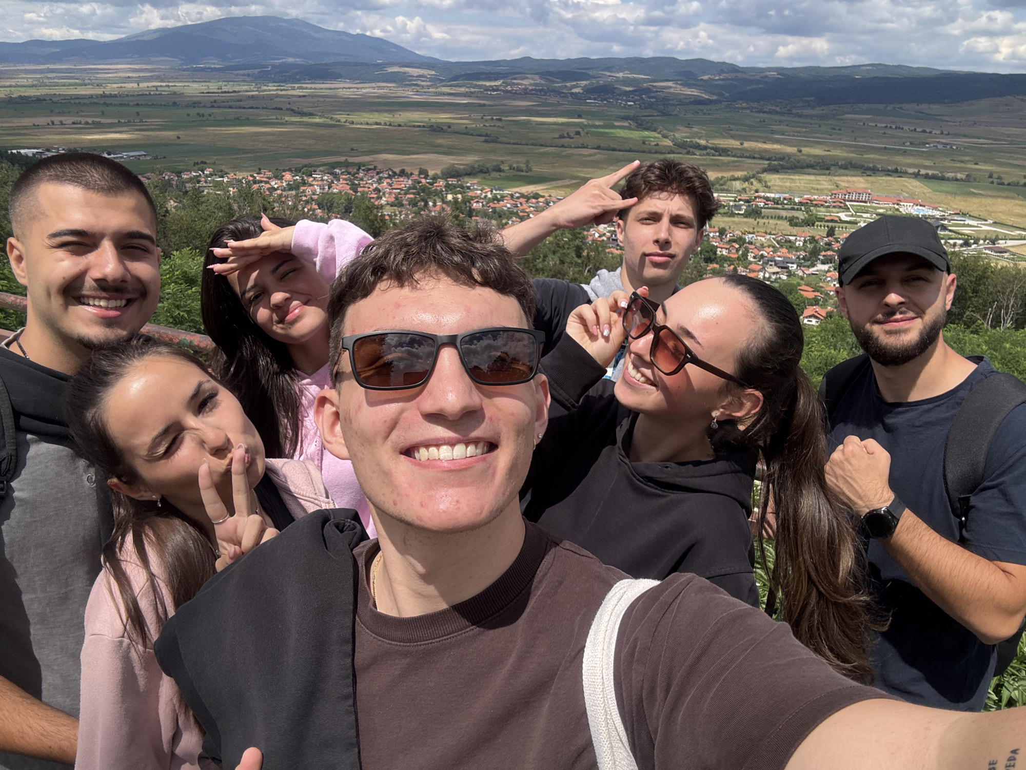 A group of seven young adults are posing for a selfie on a hilltop overlooking the town of Belchin, Bulgaria. The town is nestled in a valley with red-tiled roofs and surrounded by green fields and distant mountains. The sky is partly cloudy.
In the foreground, a young man in a brown t-shirt is holding the camera, smiling widely. To his left, a young woman in a pink hoodie is making a peace sign. Behind them, a young man in a black t-shirt and a black baseball cap is smiling at the camera. To his right, a young woman in a black hoodie is wearing sunglasses and making a peace sign. Another young man in a gray t-shirt is partially visible in the background, making a "rock on" gesture. The remaining two individuals are mostly obscured.
The overall mood of the image is cheerful and casual, suggesting a fun outing with friends. The background details, such as the town of Belchin and the surrounding landscape, provide context and a sense of place.