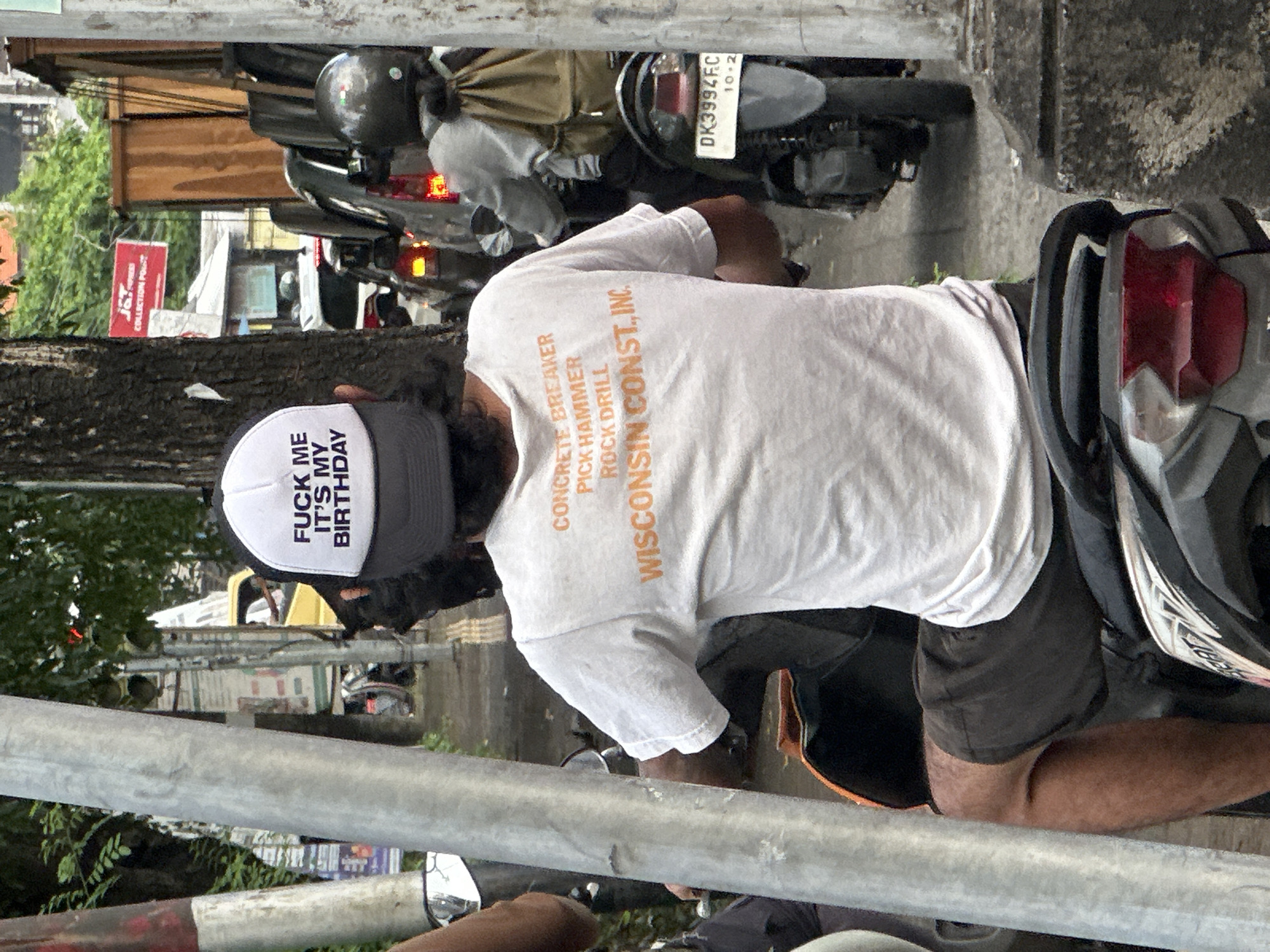 A man wearing a hat that says "FUCK ME IT'S MY BIRTHDAY" and a white T-shirt with orange text that reads "CONCRETE BREAKER, PICK HAMMER, ROCK DRILL, WISCONSIN CONST., INC." is sitting on a motorcycle. The motorcycle's license plate reads "DK3994FC" and "10-2". The motorcycle is parked on a street in Denpasar, Indonesia, near a J&T Express office. Other motorcycles are parked nearby. The environment includes trees, buildings, and metal poles.