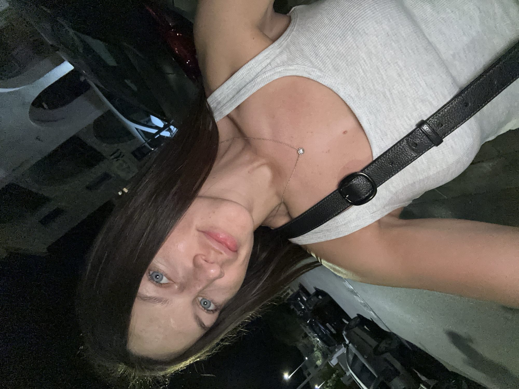 A young woman with long dark hair and blue eyes is taking a selfie at night in Budva, Montenegro. She is wearing a light grey tank top and a black leather crossbody bag. She has a delicate necklace with a small pendant. Behind her, parked cars and buildings with arched windows are visible. The streetlights cast a dim glow on the scene.