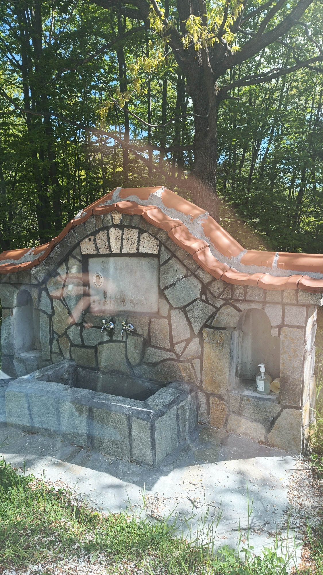 A stone fountain stands in a wooded area. The fountain is constructed of various gray stones and capped with red tiles in an arched design. Two metal faucets are visible near a plaque on the front of the fountain. A stone basin sits below the faucets. To the side, a dispenser and cup are placed on a ledge. The ground in front of the fountain is concrete, bordered by grass. Trees form a dense background, letting light through the leaves. The location is likely Vrata, Bulgaria, based on the language on the plaque.