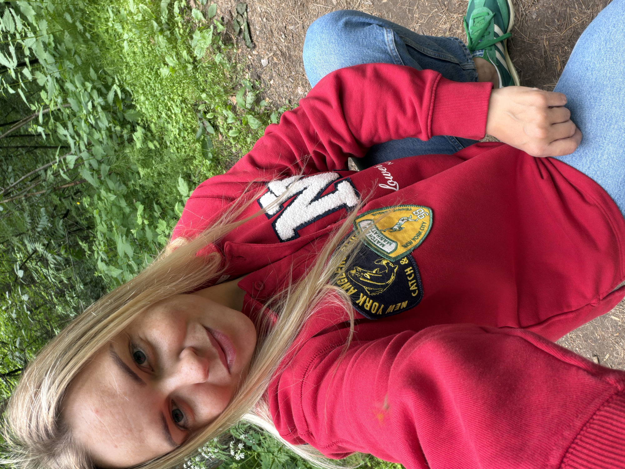 In Votkinsk, Russia, a young Caucasian woman with blonde hair, wearing a red hoodie with a large white "NY" applique and two circular patches, and light blue jeans, is captured in a close-up, high-angle selfie. She is outdoors, with lush green foliage in the background, suggesting a wooded or park-like setting. The woman is squatting, with her left hand holding the hem of her hoodie. Her facial expression is relaxed and friendly, with a slight smile. The lighting is natural, indicating daylight. The image focuses on the woman and her attire, with the natural environment serving as a backdrop.