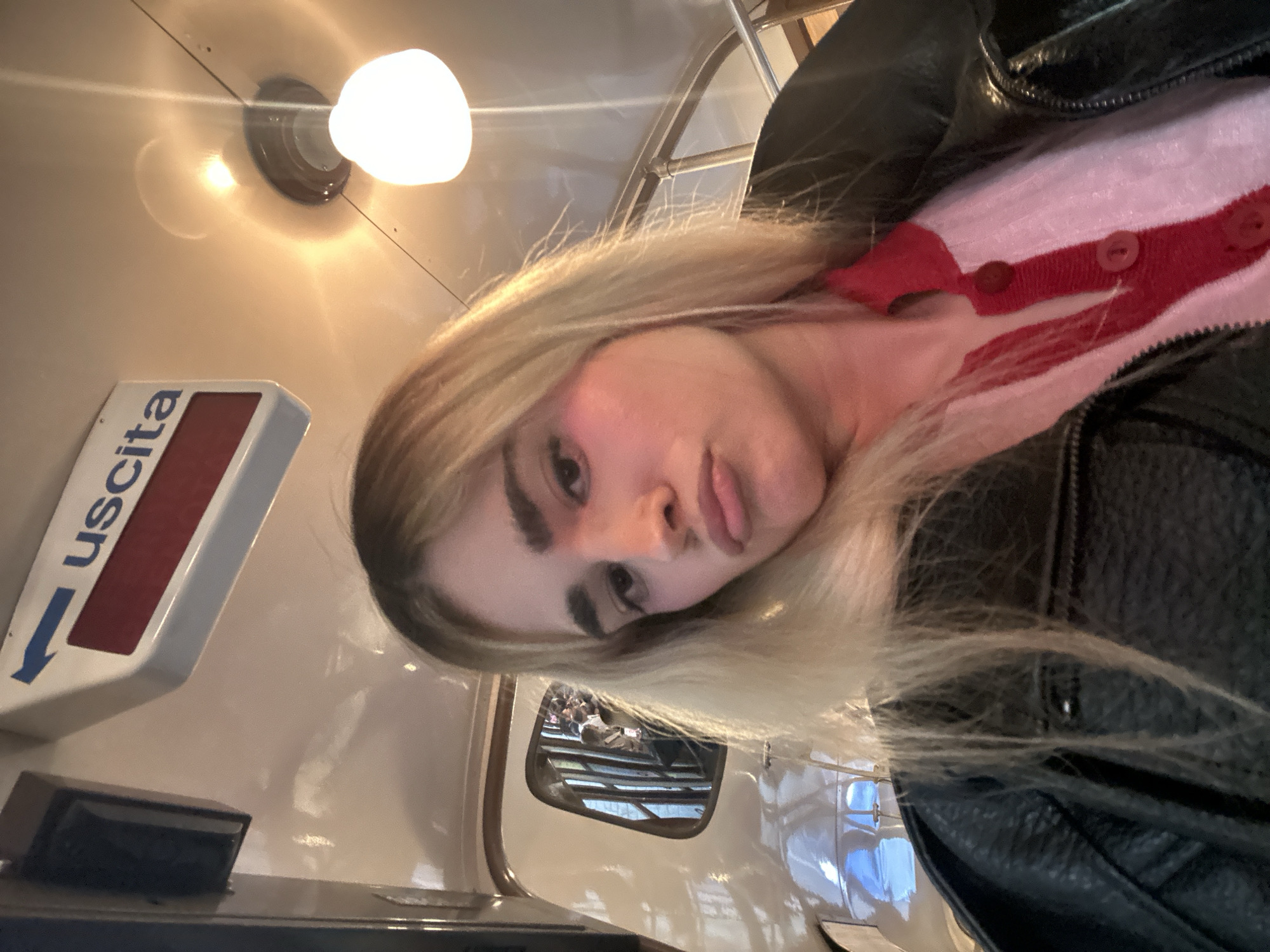 A blonde-haired woman takes a selfie while riding a historic tram in Milan, Italy. She wears a black leather jacket over a pink and red cardigan. The tram's interior is visible, including the "uscita" (exit) sign in Italian and a small window reflecting passengers and city views. A light fixture is on the tram's ceiling.
