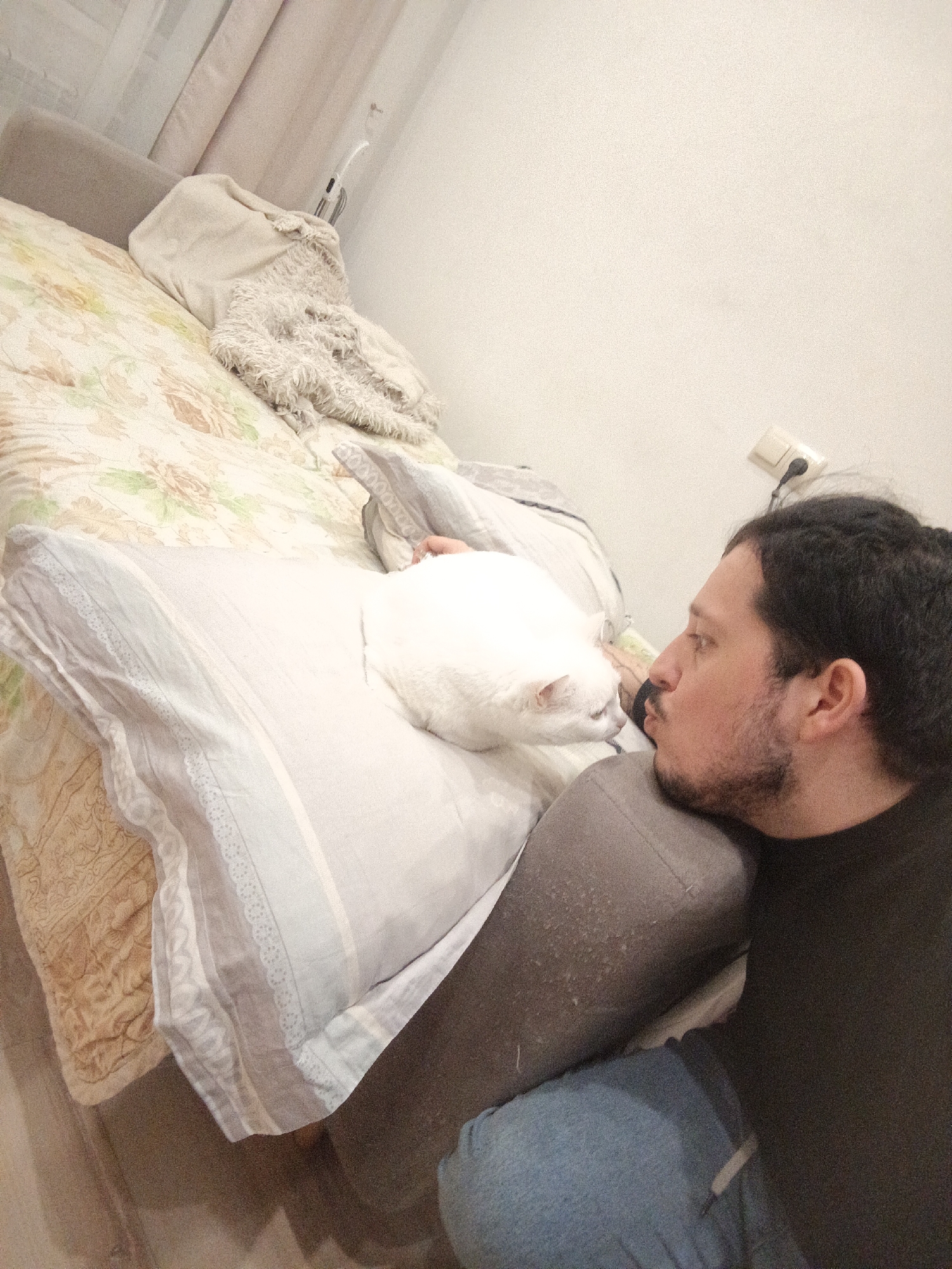 A man with dark hair, a beard, and wearing a black t-shirt and blue jeans is lying next to a white cat on a bed with light-colored linens. The bed is against a white wall with an electrical outlet. The photo was taken in Penza, Russia.