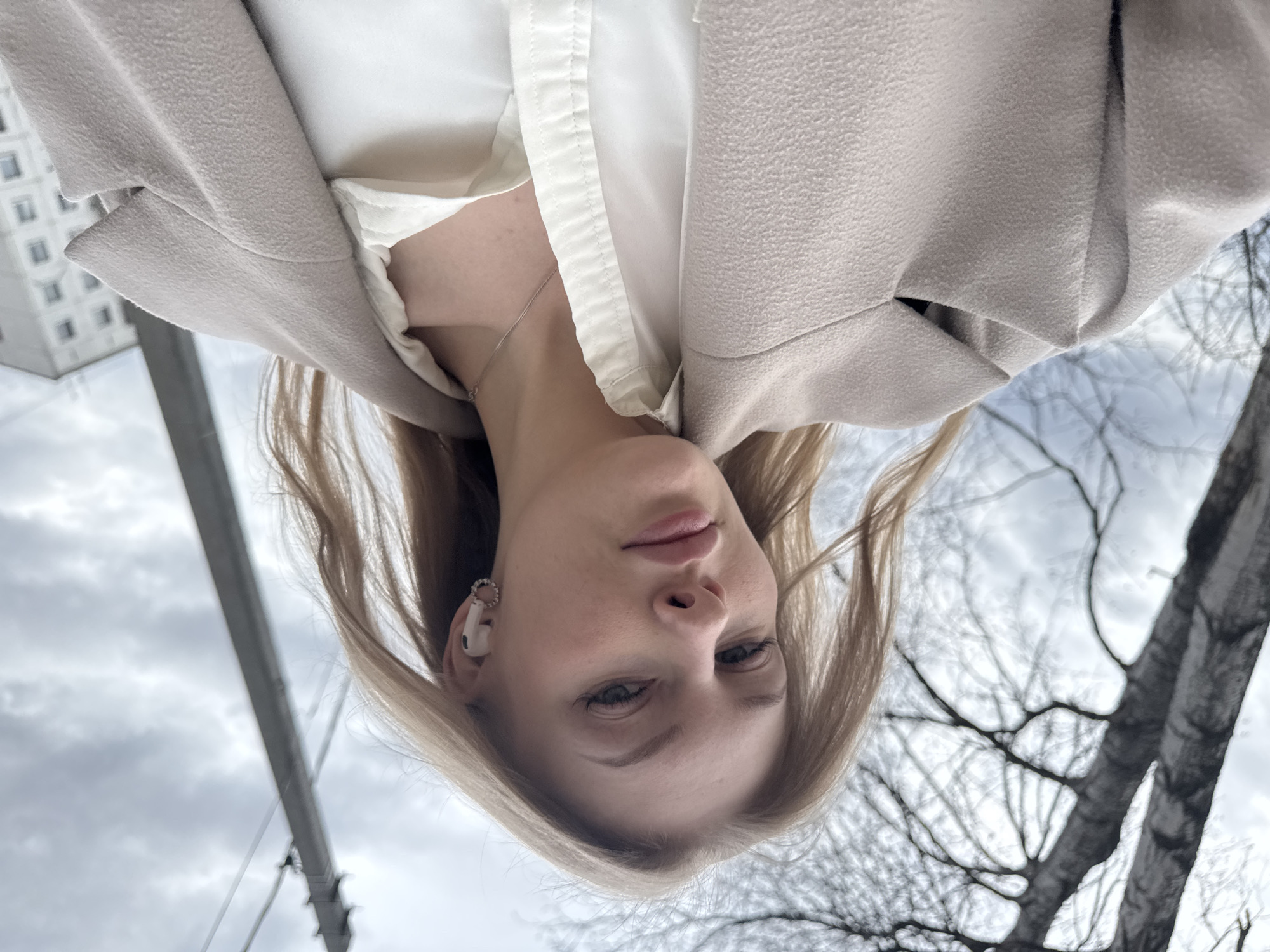 An upside-down, close-up selfie captures a young woman looking directly at the camera in an outdoor urban setting in Perm, Russia. She has light-colored hair parted down the middle and a neutral expression. Her lips are slightly parted, and her eyes appear light-colored.
She is wearing a light-colored, possibly white or off-white, collared shirt under a light beige or greyish-beige textured coat. A thin silver necklace is visible around her neck. In her left ear, she wears a white earbud, likely an Apple AirPod, accompanied by a small, decorative, circular earring or ear cuff.
The background shows an overcast sky, suggesting a cloudy day. To the top left, a modern, light-colored building with numerous windows is partially visible. To the right, bare tree branches are silhouetted against the sky, indicating a late autumn or winter season. A dark, vertical pole or structure, possibly a utility pole, is also present. The overall lighting is soft and diffused. There is no visible text in the image.