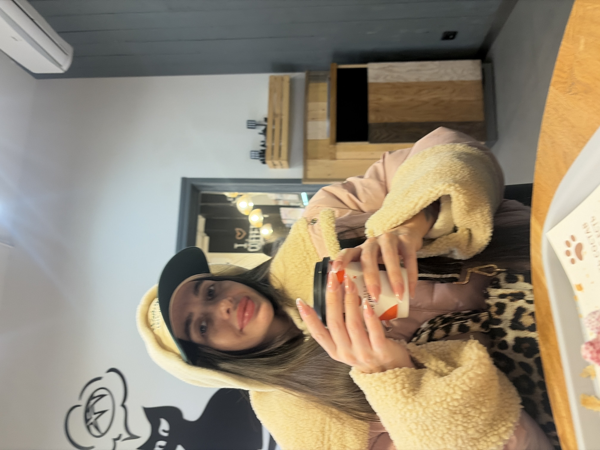 A young woman is seated at a light-colored wooden table in an indoor setting, likely a cafe, in Saint Petersburg, Russia. She looks directly at the camera, holding a white paper coffee cup with an orange design and a black lid in both hands. She wears a beige furry hooded jacket with pink quilted sections and a beige baseball cap with a dark green brim underneath the hood. Her long, dark hair is visible, and she has manicured nails. A leopard print bag strap is visible across her chest. On the table in front of her is a gray tray with white paper, featuring a paw print logo and Russian text including "СОСТАВ" (composition), alongside remnants of food items with red toppings.
The background features a white wall on the left with a large black mural depicting a stylized cat silhouette with a speech bubble containing an abstract symbol, resembling a paw print. To the right, a dark gray paneled wall is visible, with a white air conditioning unit mounted high. Below the air conditioner, a small wooden shelf holds dark decorative objects. Further right and lower, a vertical structure composed of varied wooden panels features a black rectangular opening. A dark-framed doorway in the mid-left background reveals warm-toned pendant lights and a partially visible "I LOVE COFFEE" sign, indicating an adjacent room. The decor, particularly the paw print logo and Russian text, identifies the location as a "КофеЛапа" (CoffeeLapa) cafe.