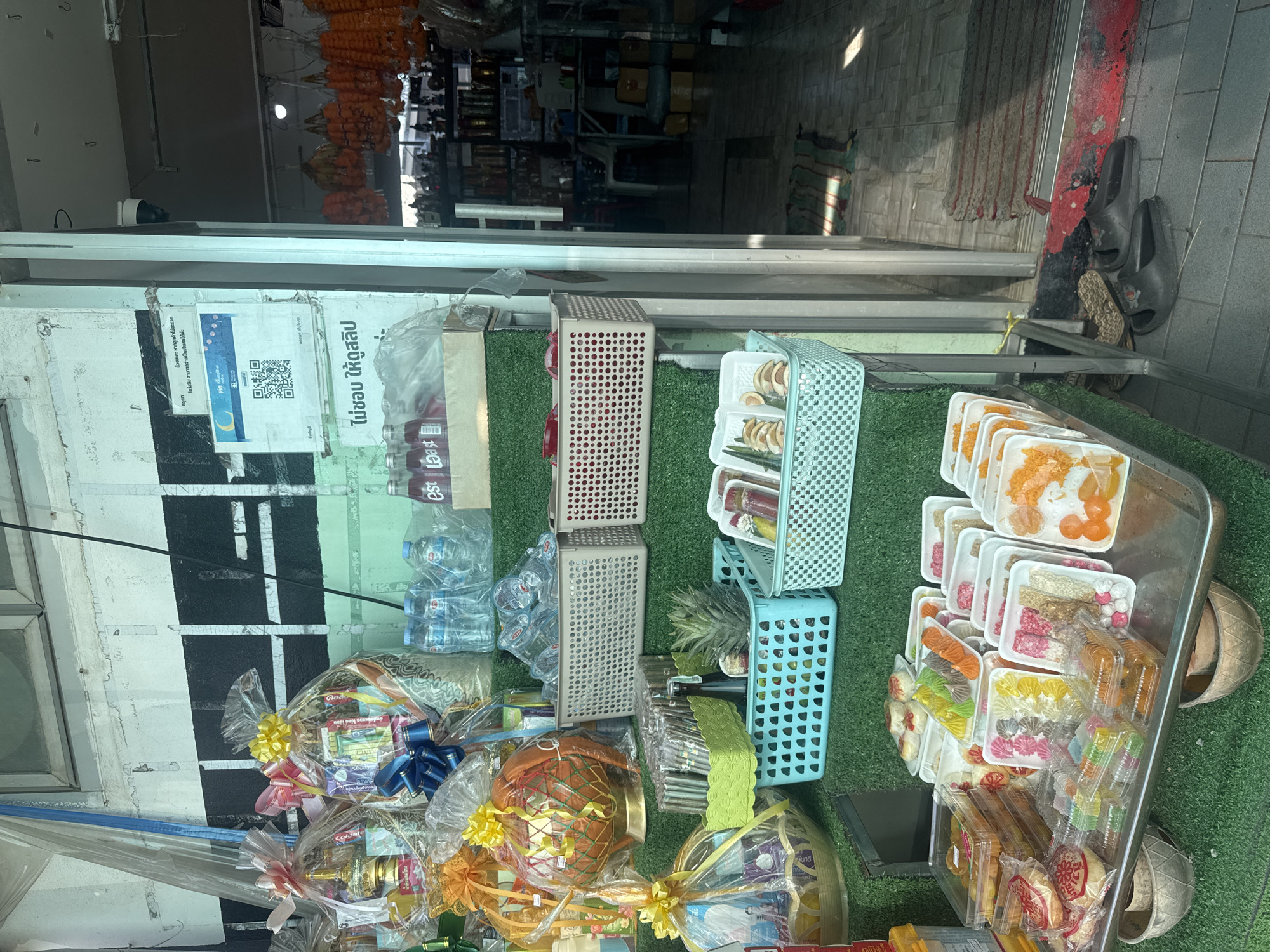 The image shows the exterior of a shop with a variety of goods displayed on shelves covered with artificial grass. The items for sale include gift baskets wrapped in cellophane, bottles of water and beverages, packaged snacks, and what appear to be traditional sweets. In the foreground, a metal tray holds numerous colorful, individually packaged desserts. A pineapple and a bottle are visible in a blue basket. Behind the displayed items, the shop interior is visible, with shelves stocked with more merchandise and a security camera mounted on the ceiling. Several pairs of shoes are placed on the tiled floor near the entrance. The overall setting suggests a small, local shop, likely in a street market or commercial area. The image is taken from a slightly low angle, looking towards the shop entrance.