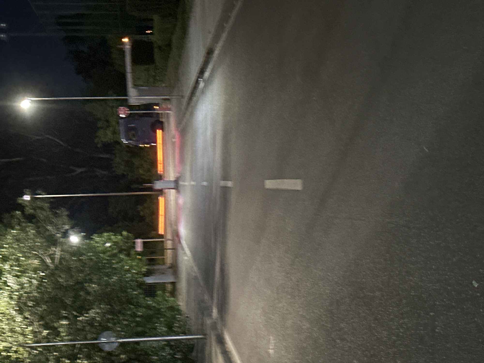 The image shows an asphalt road at night, leading towards a barrier gate. There is a blue van stopped at the gate, and the gate itself appears to be lowered, possibly blocking the road. Orange lights are visible across the road in front of the van. Streetlights illuminate the scene, casting long shadows. Trees are present on the left side of the road, and some greenery is visible on the right. The road has white lane markings. This appears to be an entrance or exit to a property, possibly a parking lot or a secured area. The image does not provide specific details that would pinpoint the location to Sofia, Bulgaria.