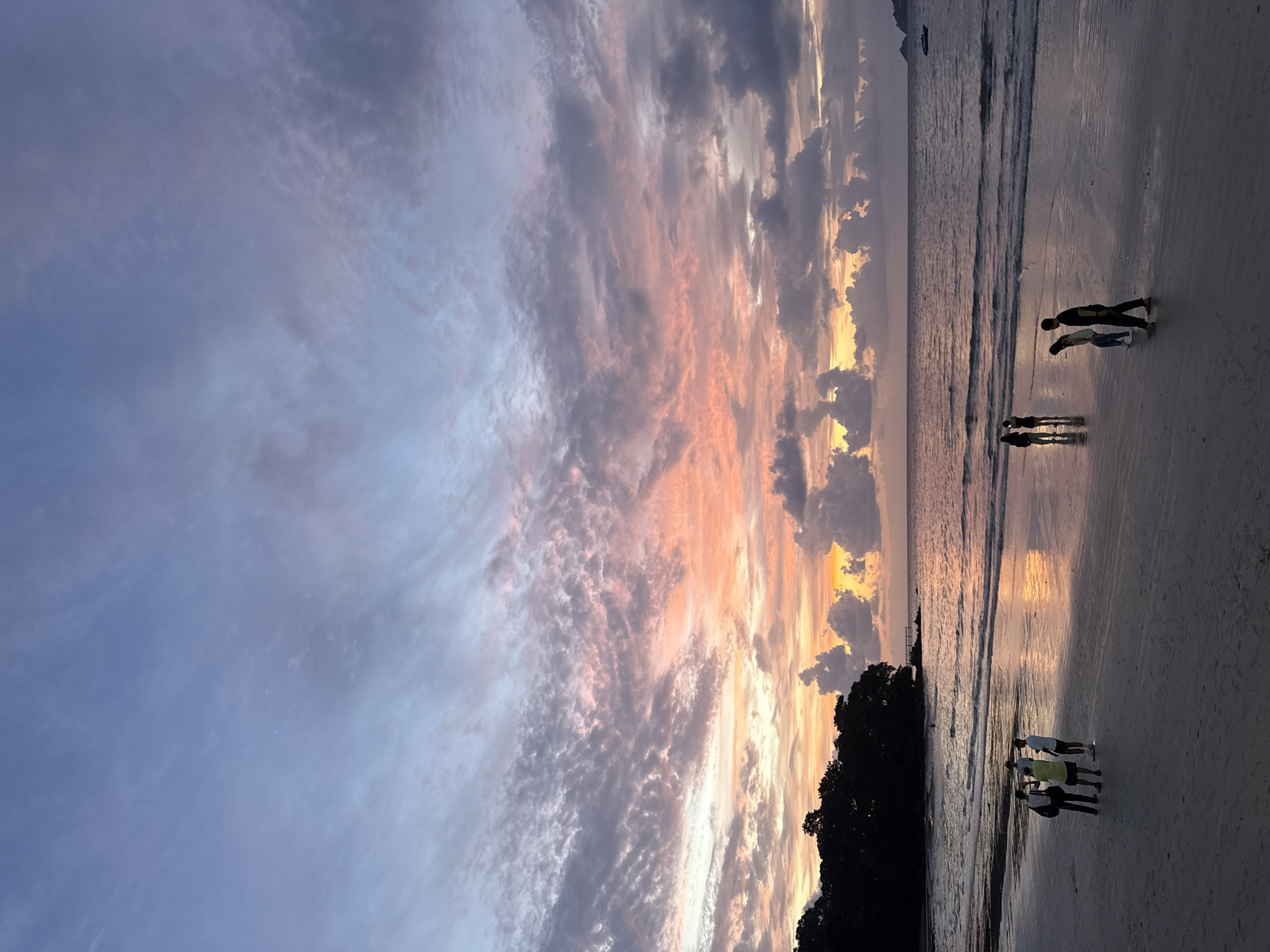 At sunset on a sandy beach in Patong, Thailand, a group of people are walking along the shore. The sky is filled with colorful clouds, painted in hues of pink, orange, and purple, reflecting on the calm ocean. In the distance, a small boat can be seen on the water. The dark silhouette of trees lines the left side of the frame, adding depth to the scene. The wet sand reflects the sky and the people, creating a shimmering effect.