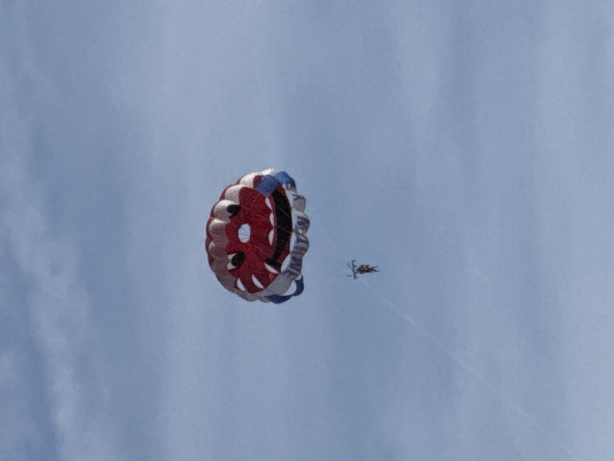 A parasailer is suspended in a bright blue sky with some wispy clouds, beneath a large, red and white parachute designed to resemble a cartoon monster face. The parachute has a wide, grinning mouth with sharp white teeth, two black eyes, and a red face with white accents. The parasailer, a small figure in the distance, is attached to the parachute by lines. The scene is observed from below, looking upward at the sky. The location is identified as Budva, Montenegro.