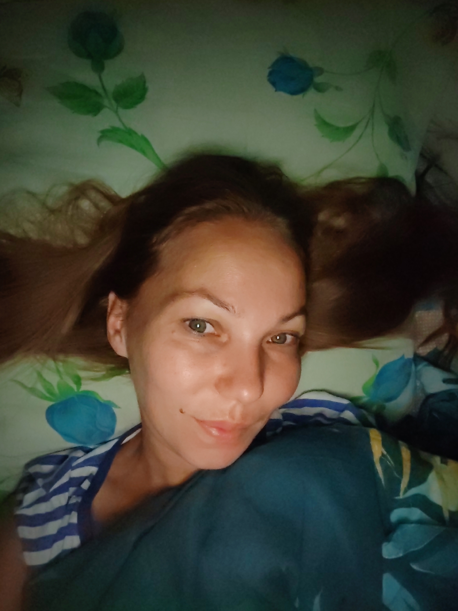 The image is a close-up selfie of a woman lying on her back, with her head on a pillow. The pillow has a light green background with a blue rose pattern. The woman has long, light brown hair spread out around her head. She is wearing a blue and white striped t-shirt and a dark teal blanket is covering her lower body. Her face is visible, with her eyes open and looking at the camera. She has fair skin and a mole on her left cheek. The lighting is dim, creating a soft glow on her face. There is no visible indication of the location within the city of Чухлэм, Russia.