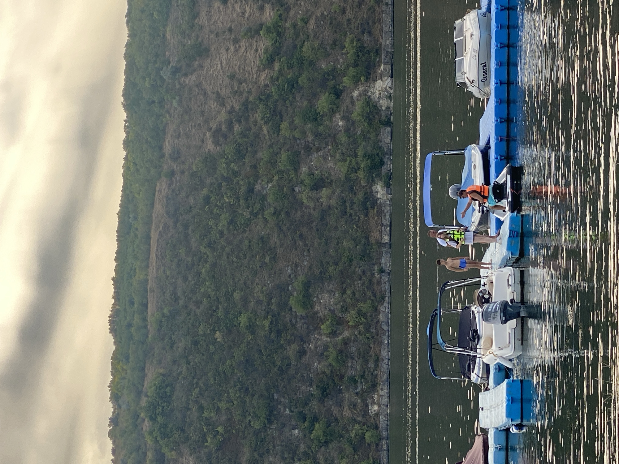 In Molovata Nouă, Moldova, a body of water, likely the Nistru River, is calm with ripples reflecting the overcast sky and the lush green hillside that slopes down to the water's edge. A floating dock system, composed of blue modular plastic floats, extends from the left side of the frame. Several boats are moored to the dock.
On the dock, near the center, are two children and two adults. A young boy in blue swim trunks stands next to a girl wearing a life jacket and white shorts, who is holding a rope. Behind them, a woman in a white top and shorts also wears a life jacket. To the right, a man in an orange life jacket and turquoise shorts rides a jet ski. He is gesturing with his left hand.
In the background, docked to the floating platform, are several watercraft. The closest boat to the left has a dark cover and a blue canopy. Next to it is a white motorboat with a light brown interior and a Yamaha outboard motor. To the right of this is a boat with a blue canopy and a white hull. Further to the right, a white boat with the word "General" visible on its side is also docked. The landscape behind the water is dominated by a steep, tree-covered hill.