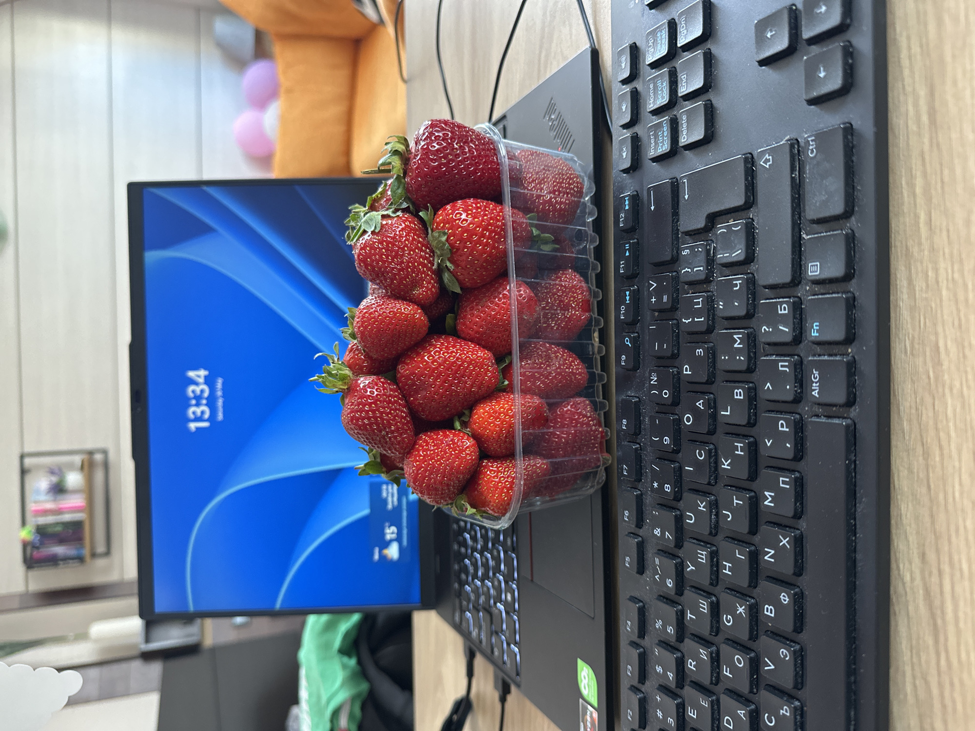 A desk scene shows a monitor displaying a time of 13:34 and a temperature of 15 degrees Celsius, positioned next to a laptop with a container of strawberries placed on top. A black keyboard sits to the right of the laptop. The room features a wall with shelves and decorations in the background. The image does not provide enough information to determine the location in Varna, Bulgaria.
