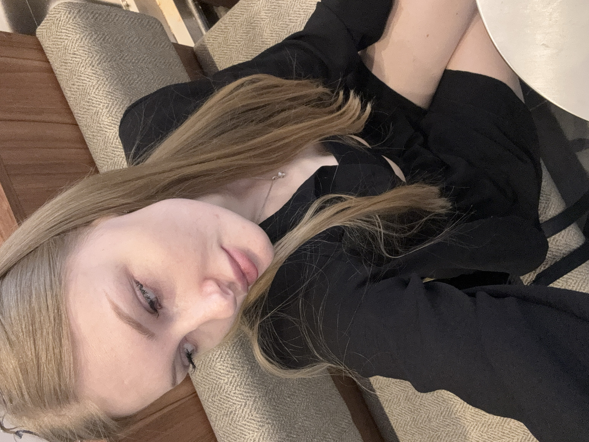 A woman with long blonde hair is lying on a beige upholstered chair. She is wearing a black blazer and a necklace. The chair is positioned near a wooden surface. Next to her is a white plate, possibly on a table. The location is suggested to be Perm, Russia, though no specific landmarks are visible in the image to confirm this.