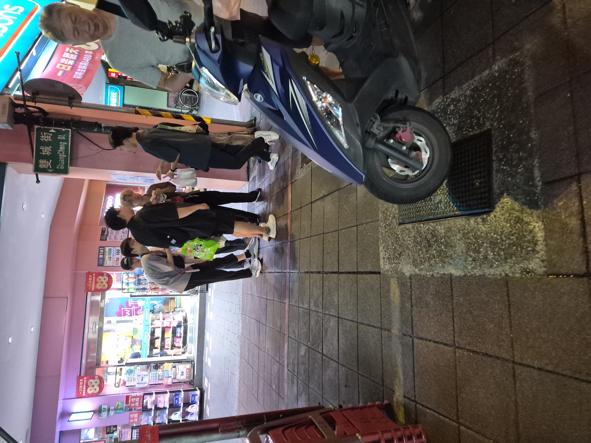 The image captures a street scene in Taipei, Taiwan, specifically at the intersection of Shuangcheng Street. In the foreground, a blue and purple scooter is parked on a sidewalk composed of square paving stones. The scooter's front wheel is positioned over a metal grate.
To the left, a group of four young men are gathered. Two are wearing black shirts and pants, one is in a grey t-shirt and dark pants, and another is in a black shirt and grey pants. One man in the black shirt is holding a white plastic bag. They appear to be interacting, with some looking at each other.
Behind them, a brightly lit store with a pink facade is visible. Red banners with the number "88" are displayed. The store appears to be a convenience store or pharmacy, with shelves of products visible through the glass doors. Further down the street, other store signs and awnings can be seen.
To the right of the scooter, an older man wearing a grey t-shirt and dark pants is walking away from the camera, with a dark strap across his chest. Another person, partially visible, is standing near him. The overall lighting suggests it is evening or nighttime, with artificial lights illuminating the street and storefronts. The sidewalk is wet in places, possibly from recent rain.