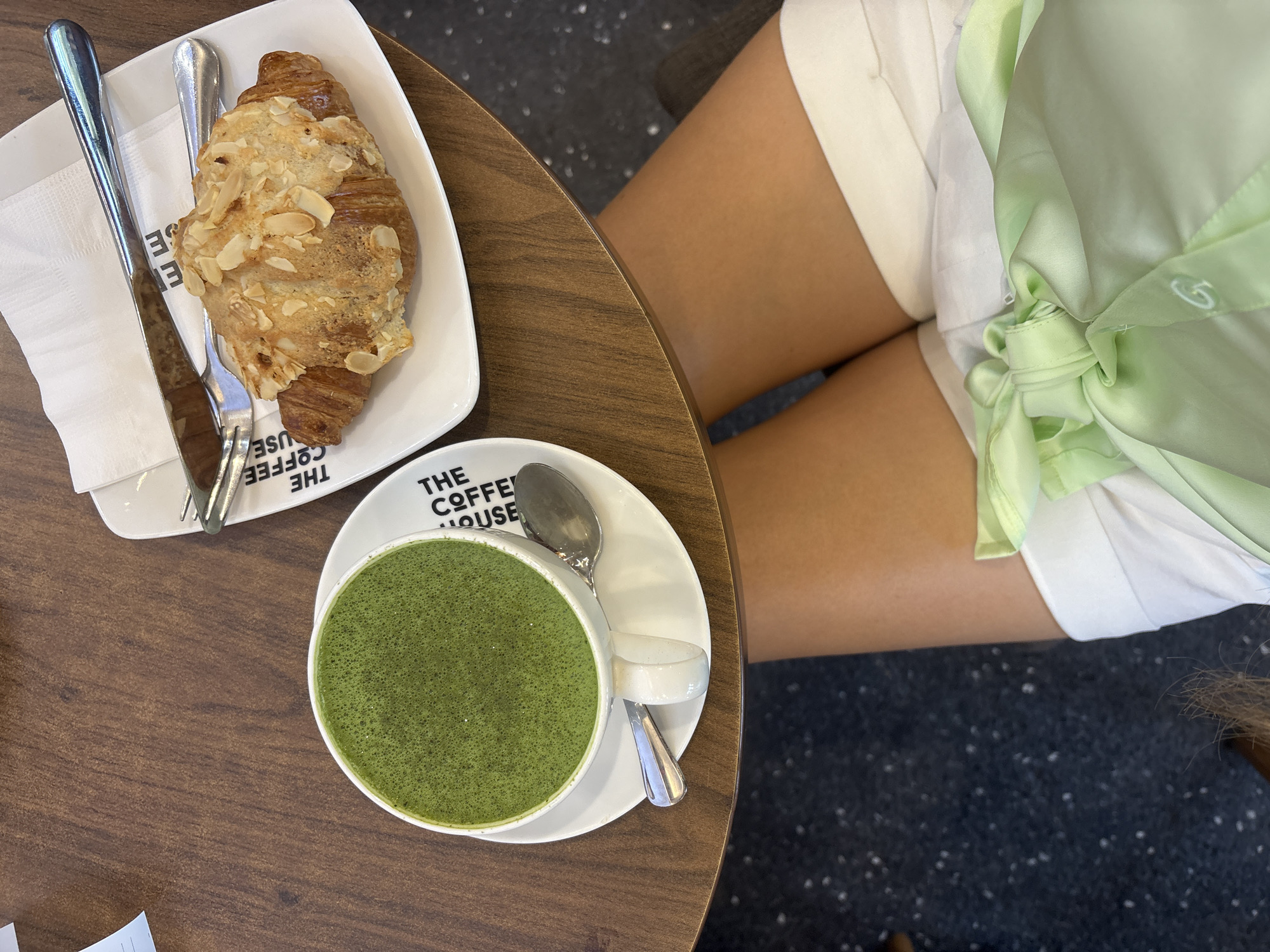 A photograph taken from above depicts a person seated at a wooden table in a coffee shop in Nha Trang, Vietnam. A white plate bearing an almond croissant and silverware sits on the table. A cup of green matcha tea rests on a saucer that also bears "THE COFFEE HOUSE" logo. The person is wearing white shorts and a light green shirt. The floor is a dark color with small white specks.
