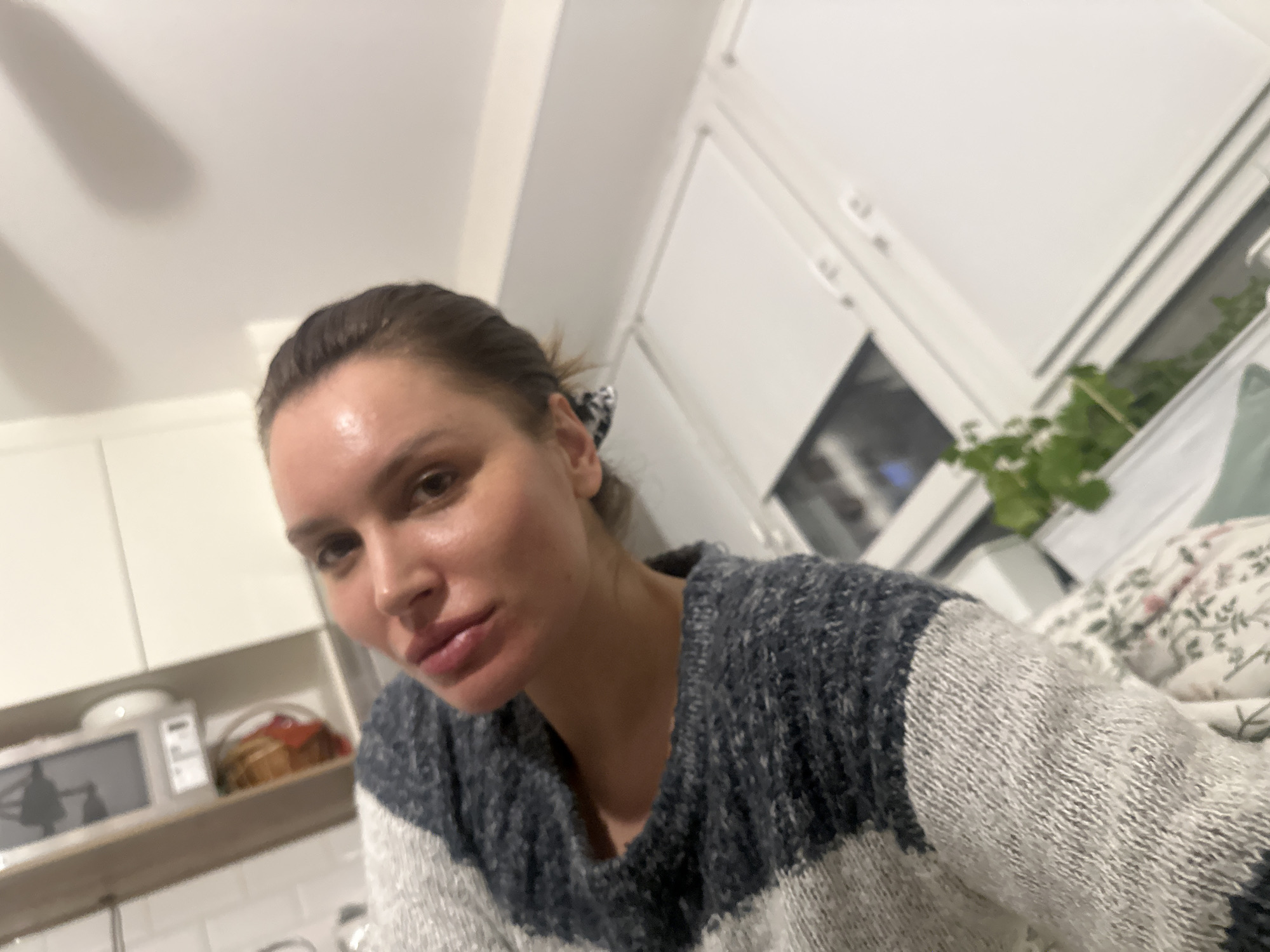 A woman is featured in a close-up indoor selfie. She is looking directly at the camera with a neutral expression. Her hair is pulled back, with a patterned scrunchie visible behind her right ear. She is wearing a thick, striped knit sweater in shades of dark grey/blue and white.
The background suggests a domestic setting, likely a home. To the left, white kitchen cabinets are visible, with a white microwave oven placed on a shelf below them. Next to the microwave, a basket containing what appears to be baked goods is present. On the right side of the frame, a window or glass door is partially visible, with green foliage from a plant situated near it. Further right, a portion of a floral-patterned pillow or blanket is seen. The lighting suggests an indoor setting with artificial illumination, possibly in the evening. There is no visible text in the image.