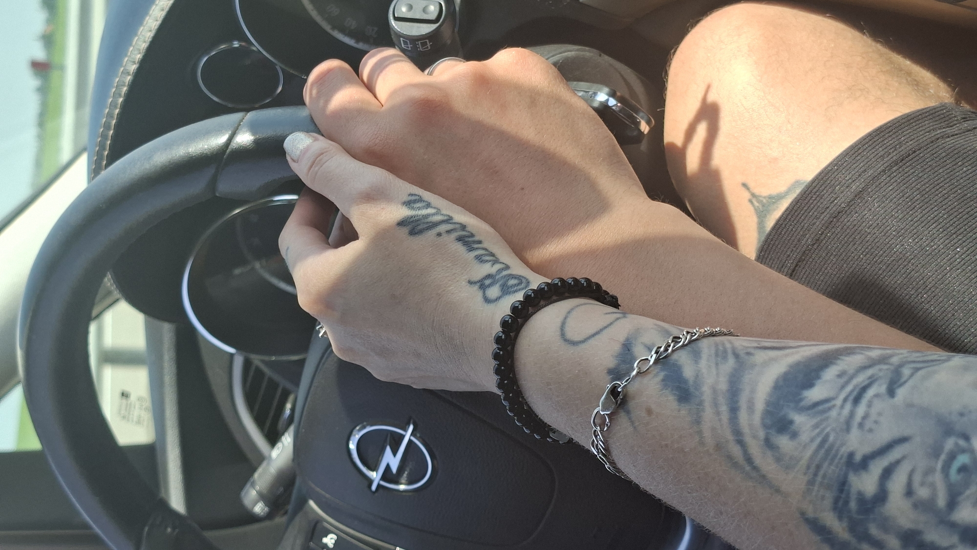 Two people are in the front seats of a car. One person's hands are on the steering wheel, and the other person's hands are resting on the first person's hands. The first person has a tattoo on their hand that says "Kamilla". The first person is also wearing a black beaded bracelet and a silver chain bracelet. The second person is wearing a ring on their finger. The car appears to be driving on a road with a grassy area and trees in the background. The image is taken from inside the car, looking forward. The location is identified as Самахвалавіцкі сельскі Савет, Belarus.