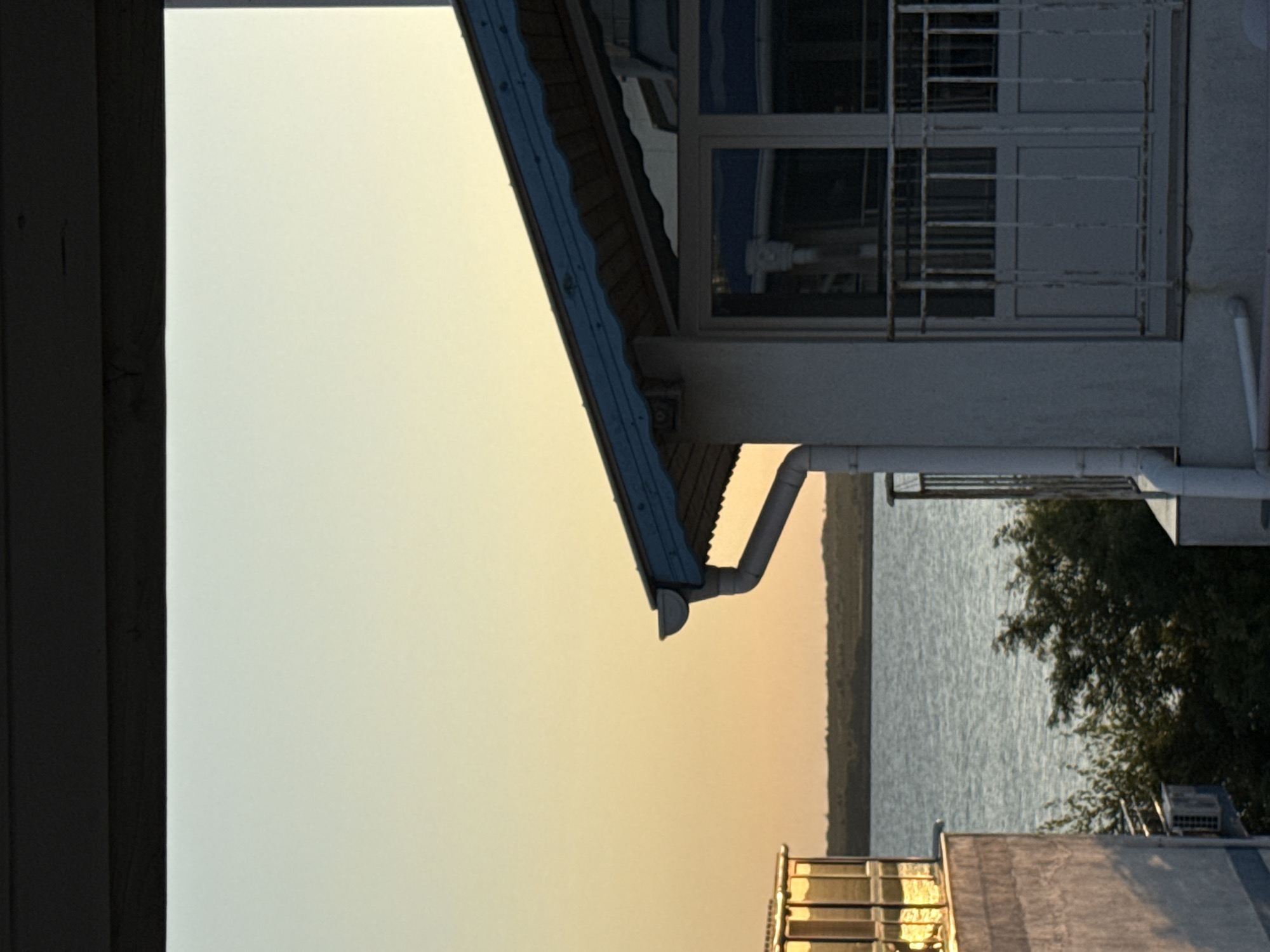 The image captures a view of the sea and buildings in Pomorie, Bulgaria, during what appears to be sunset or sunrise. In the foreground, a white building with a blue-trimmed roof and a balcony is visible. A white downspout runs down the side of the building. To the left, another building, partially obscured, features glass windows that reflect the warm light. Beyond the buildings, a body of water stretches towards a distant, low-lying landmass covered in vegetation. The sky is a soft gradient of yellow and pale blue. The overall impression is one of tranquility and coastal scenery.