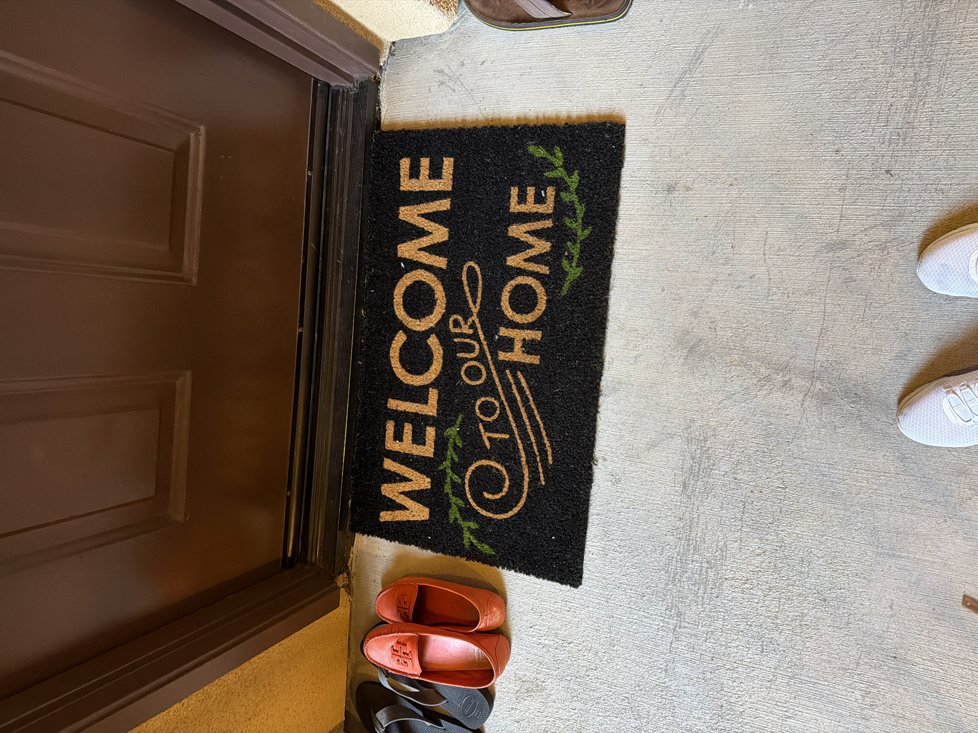 The image shows a doormat with the words "WELCOME TO OUR HOME" in gold lettering on a black background, placed in front of a dark brown door. To the left of the doormat are two orange slip-on shoes and a pair of gray flip-flops. To the right of the doormat, a brown flip-flop is partially visible. In the lower portion of the image, the toes of a person wearing white sneakers are visible. The doormat and shoes are on a concrete surface. The background consists of a textured wall, a door, and the concrete floor. The scene appears to be an entryway to a residential building.