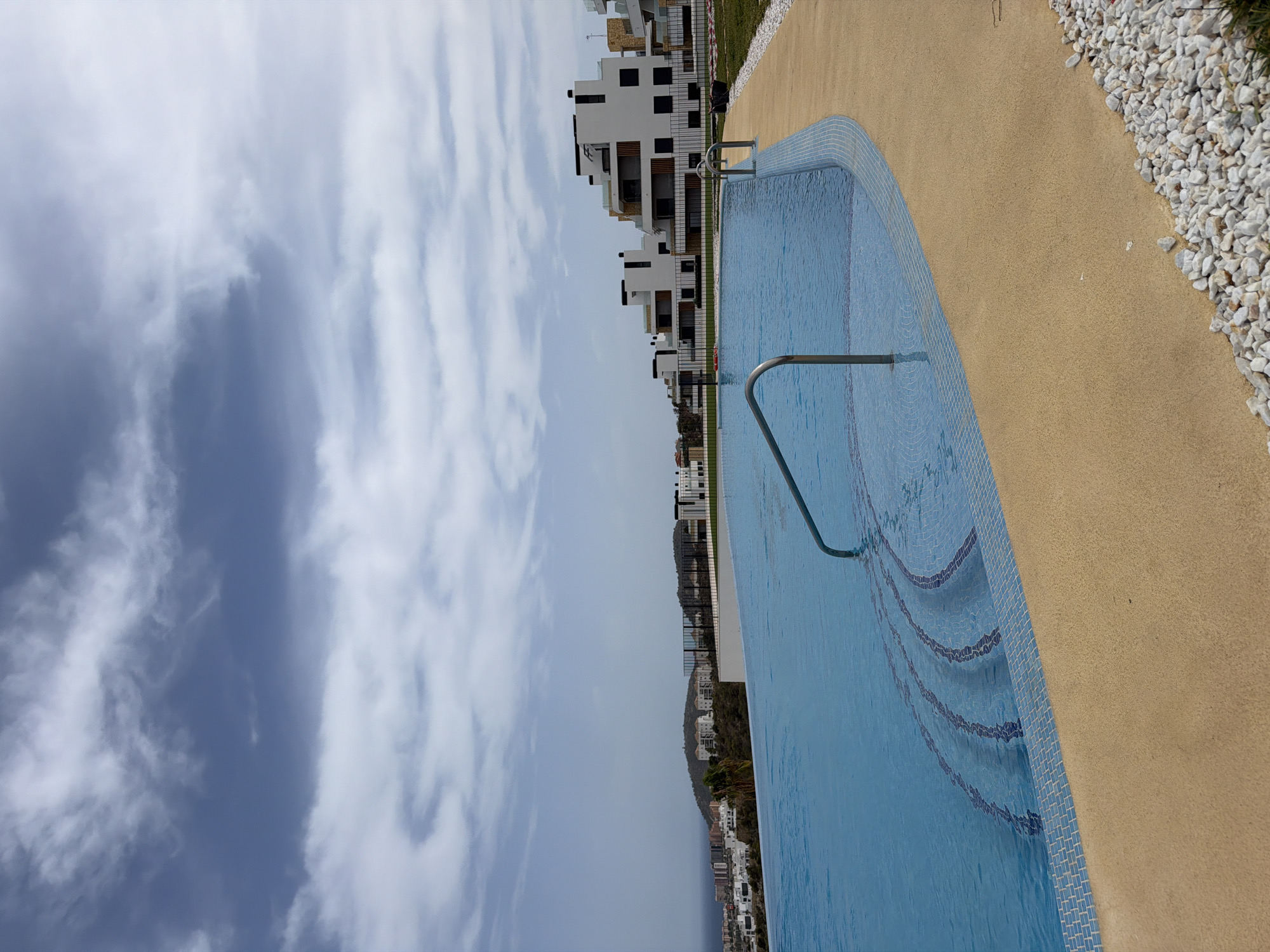 The image captures an infinity pool with a tiled edge and steps in the foreground, set against a backdrop of modern apartment buildings and a distant cityscape. The sky above is partly cloudy, with dramatic formations of white and gray clouds against a blue canvas. In the middle ground, a row of contemporary white buildings with dark window accents and balconies lines a ridge, overlooking the Mediterranean Sea and a coastal town. The architecture suggests a residential complex, possibly overlooking the town of Finestrat, Spain, given the characteristic hilly terrain and coastal proximity often seen in that region. The pool itself is a vibrant blue, reflecting the sky, and its edge is lined with small, blue and white mosaic tiles. A metallic handrail curves into the water, leading to submerged tiled steps. The area surrounding the pool features a light-colored sandy path and a border of white pebbles. The overall scene depicts a serene, upscale residential area with a focus on leisure and views.