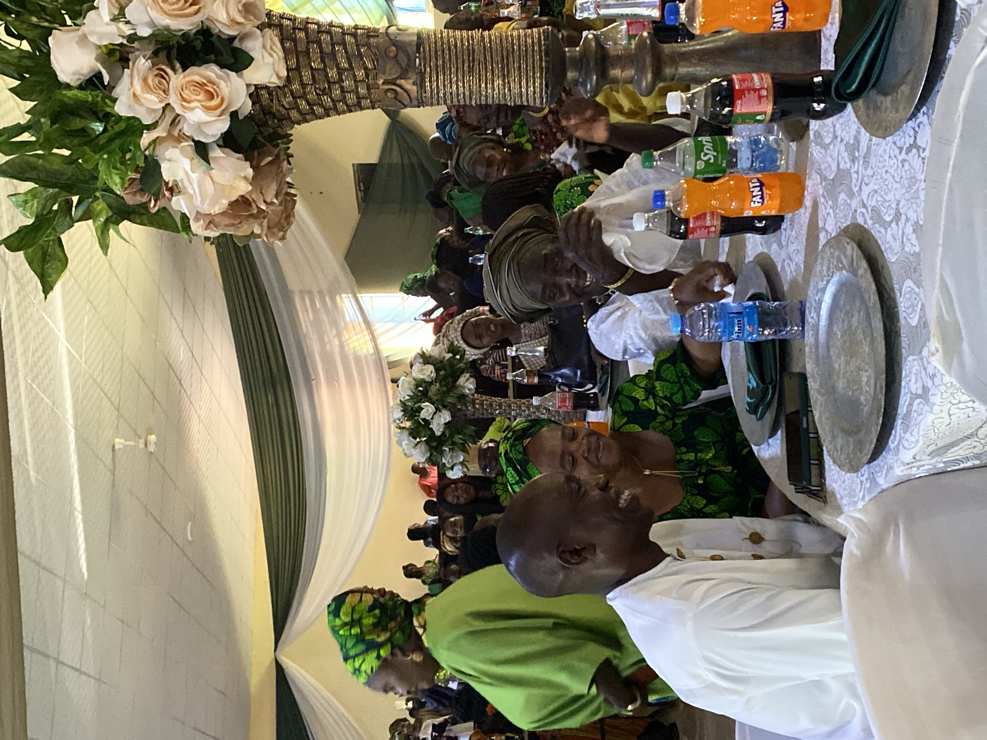 A medium, indoor shot depicts several people sitting at banquet tables set for a formal occasion, most likely a wedding, in Vom, Nigeria. The camera is positioned at an angle, revealing the left side of the tables and the individuals seated around them. The room is decorated with white and green drapery from the ceiling, and floral centerpieces adorn each table.
Many of the attendees are wearing traditional Nigerian attire, with vibrant colors and patterns. Some individuals are smiling and looking towards the camera, while others are engaged in conversation or interacting with items on the table.
The tables are covered in white tablecloths with patterned designs. Each setting includes silverware, plates, and a variety of bottled beverages like water, Coca-Cola, Sprite, and Fanta. There is a general sense of celebratory activity and merriment in the scene.