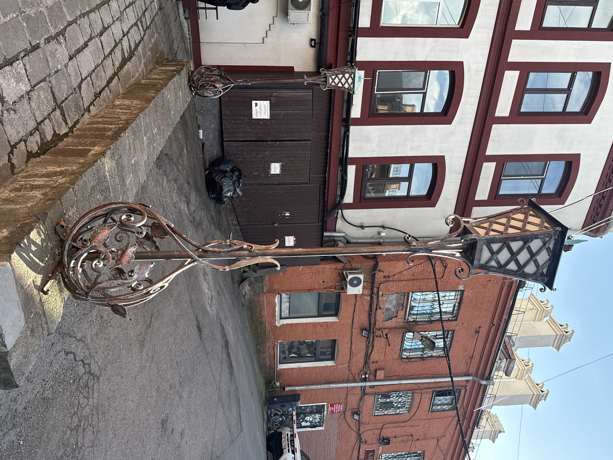 The image depicts an alleyway in Vladivostok, Russia, with two ornate wrought-iron lampposts in the foreground. The left lamppost is closer to the viewer, positioned on a stone curb. Its base features a swirling metal design with leaf motifs. The right lamppost, slightly further back and partially obscured, appears to be identical.
The alleyway is paved with asphalt, showing some cracks and wear. On the left side of the alley, a two-story building with a white facade and dark red trim surrounds multiple windows. A dark brown wooden door is visible on the ground floor, with two large black trash bags piled in front of it. To the left of the door, an air conditioning unit is mounted on the building's wall.
On the right side of the alley, a three-story brick building displays a weathered facade with several windows, some of which are barred. The upper stories have prominent white chimneys. A white SUV is parked on the right side, partially blocking a metal barrier and graffiti-covered wall. The sky above is clear and blue.