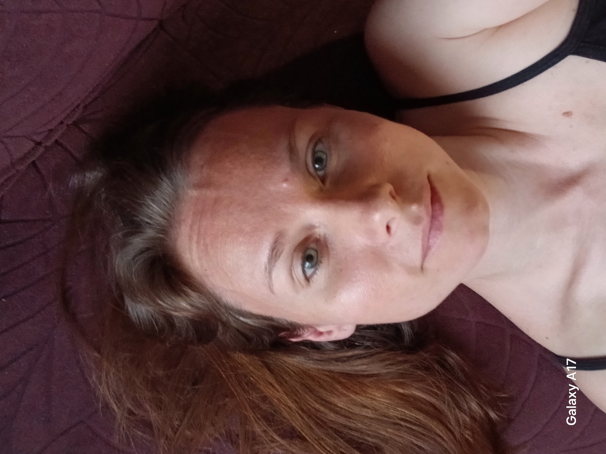 A woman with fair skin, light-colored eyes, and auburn hair lies on a burgundy-colored textured surface, possibly a bed or couch. She is wearing a black tank top. The photograph appears to have been taken with a Samsung Galaxy A17 phone, judging by the watermark in the bottom right corner. The location in Yekaterinburg, Russia is unknown.