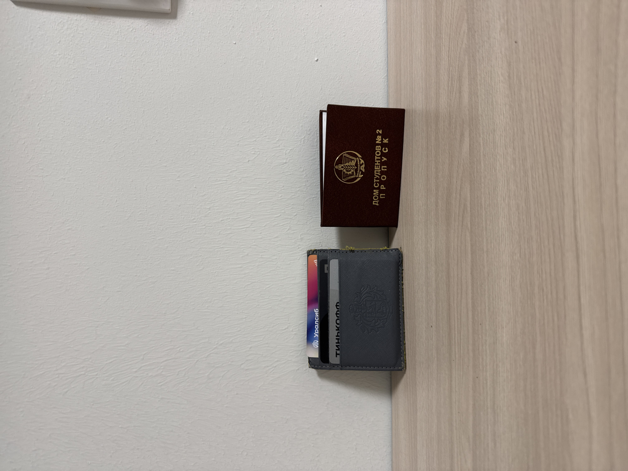 A close-up shot shows a brown document with gold lettering and a grey wallet with credit cards, placed on a surface where a white wall meets a light wood-grain door. The lettering on the brown document reads "ДОМ СТУДЕНТОВ №2 ПРОПУСК". The wallet displays cards and a logo. The image suggests an interior setting. The language on the document and cards indicates a location in Russia. Specific mention of "ДОМ СТУДЕНТОВ" (student residence) and the bank names "Уралсиб" and "Тинькофф" suggest a location in Kazan.