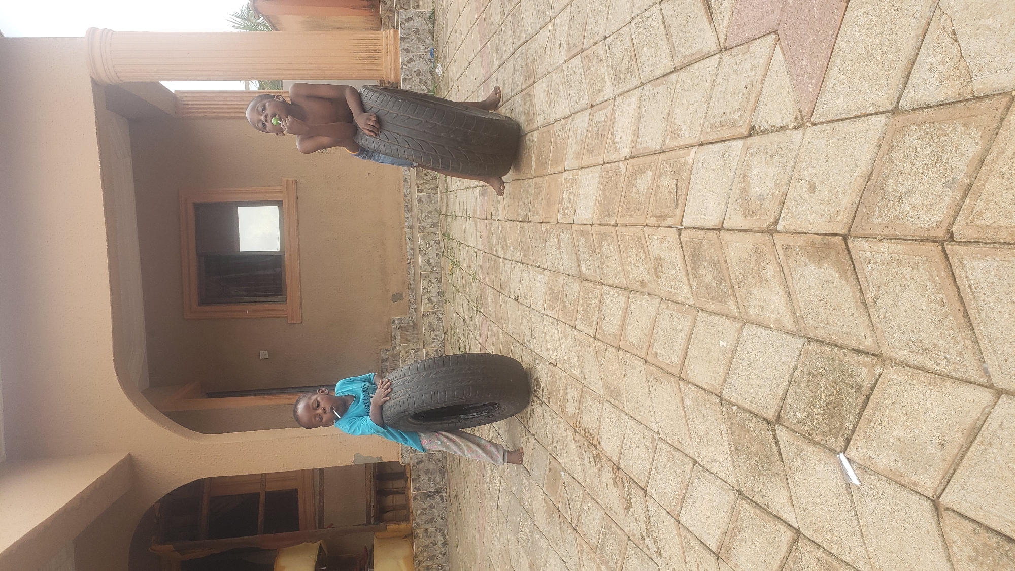In Sabon Tasha, Nigeria, two young boys are playing on a paved courtyard in front of a beige, two-story building. The building features arched doorways, windows with light-colored frames, and a prominent fluted column on the right side.
The boy on the left, wearing a light blue t-shirt with "MONSTER" visible and patterned pants, stands barefoot beside a large, black tire. He is holding a white lollipop to his mouth.
The boy on the right, shirtless and wearing blue shorts, also stands barefoot next to a similar large, black tire. He is leaning on the tire and also appears to be holding something to his mouth.
The courtyard is paved with light-colored square and diamond-shaped stones. The lighting suggests it is daytime.