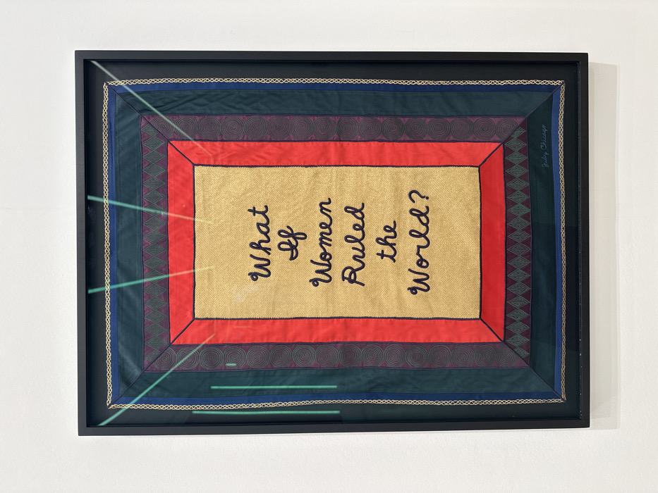A framed textile artwork, likely an embroidered quilt or tapestry, is centrally displayed against a plain white wall, suggesting a gallery or museum environment. The artwork, oriented vertically, features a black frame with a reflective glass or acrylic cover, exhibiting several faint green linear reflections from ambient lighting.
The artwork itself is rectangular, with a central golden-colored panel containing black cursive text that reads, "What If Women Ruled the World?" This central panel is bordered by a solid red fabric strip. Beyond the red border are multiple concentric borders made of dark green and dark blue fabrics, incorporating various embroidered patterns. These patterns include repeating circular spirals on vertical strips, and a horizontal band with interlocking diamond or triangular motifs. The signature "Judy Chicago" is visible in small, black script in the bottom right corner of the innermost dark green border, just above the red border. No individuals are present in the image. The specific location within the city of London, United Kingdom, cannot be determined from the provided visual information.