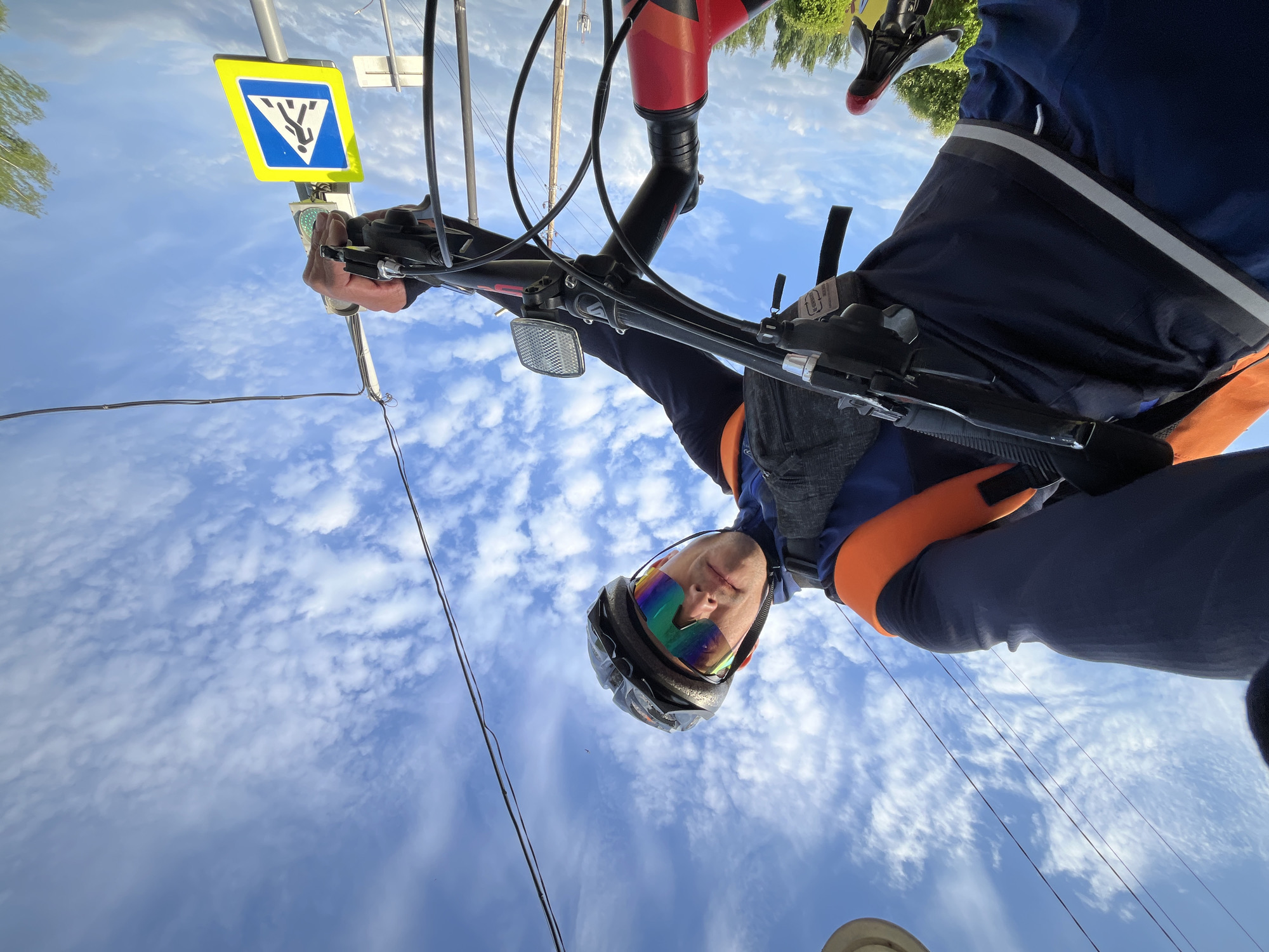 A person is riding a bicycle beneath a blue sky filled with clouds in Chaikovsky, Russia. The person is wearing a helmet and glasses. In the background is a pedestrian crosswalk sign.