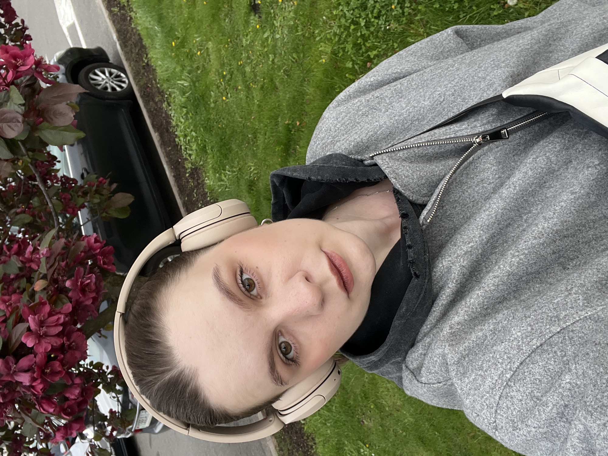 A person wearing headphones and a gray hooded jacket is shown in a selfie-style image, seemingly taken outdoors. The background includes green grass, a tree with red-purple flowers, and a car parked along what appears to be a street. The location cannot be verified from the image as Minsk, Belarus.