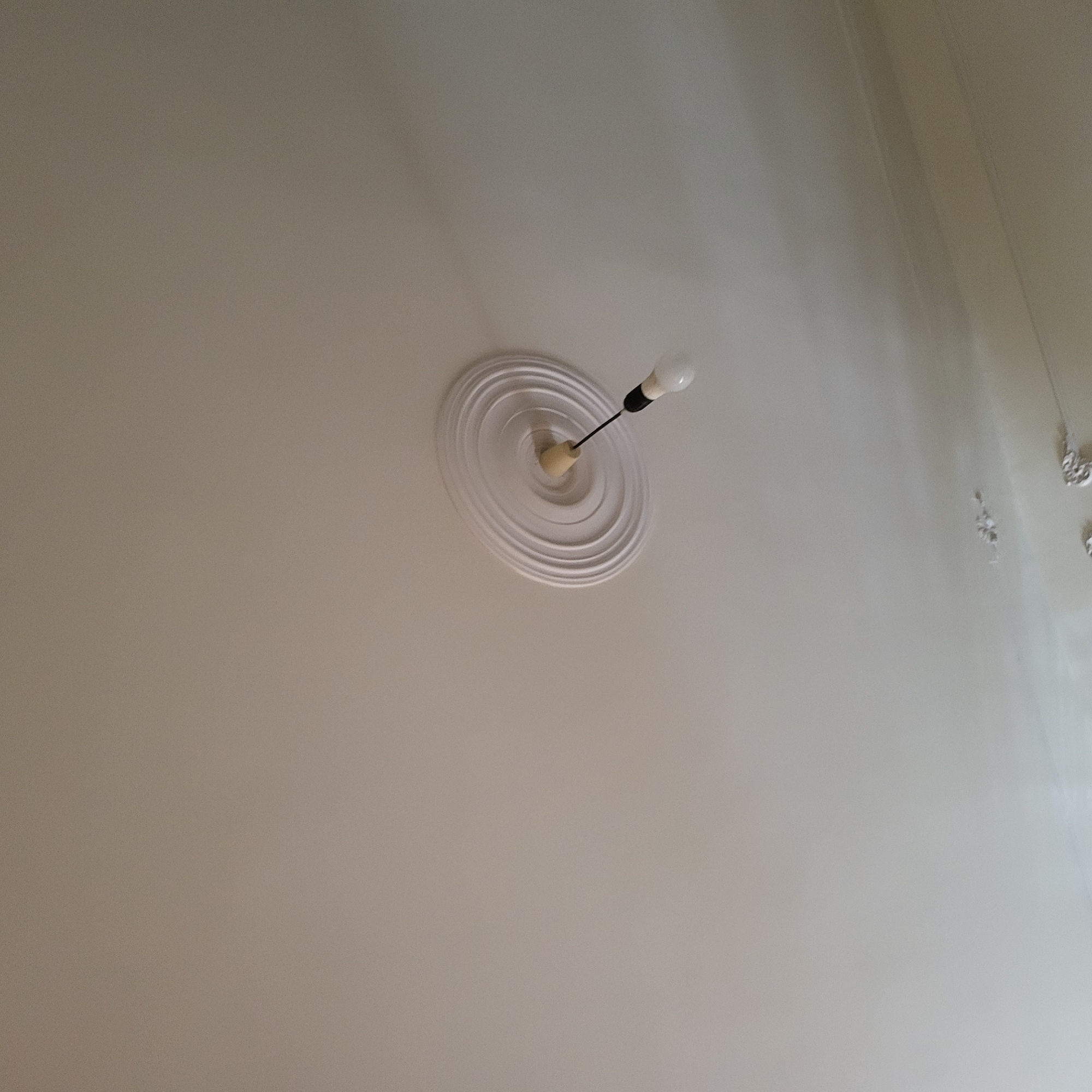 The image displays a light-colored ceiling, appearing to be off-white or a very light grey, viewed from below and at an angle. The primary subject is a simple light fixture situated towards the upper center of the frame. This fixture consists of a single bare lightbulb hanging from a dark electrical wire. The wire emerges from a small, light-colored, possibly wooden, cap, which is centrally positioned on a decorative, circular ceiling rose. The ceiling rose features several concentric, raised rings, adding a classical architectural detail to the ceiling.
On the far right side of the ceiling, several faint, intricate decorative elements are visible, potentially plasterwork or remnants of another fixture, suggesting a more elaborate ceiling design in that area. A subtle, dark, linear mark or shadow also runs vertically along the right edge of the frame. The overall surface of the ceiling shows minor variations in tone, indicating uneven lighting or slight textural differences. No people or activities are present. There is no visible text. The setting is an indoor space, likely a room in a building in Yerevan, Armenia.