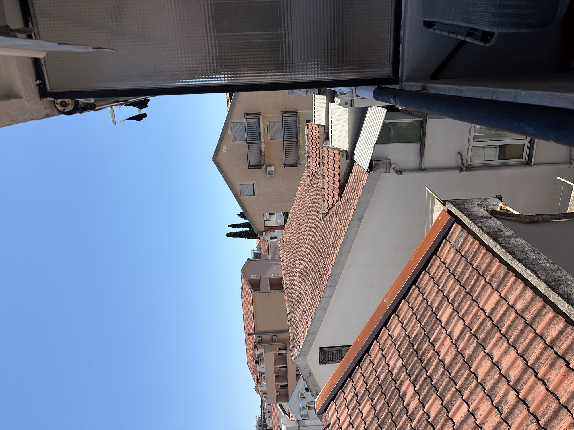 A slightly rotated view from a window in Budva, Montenegro, looking out at neighboring buildings under a clear blue sky. The buildings feature terracotta tile roofs, white or beige facades, and some have air conditioning units mounted on their exteriors. From the vantage point, details of the window, including the textured glass and frame, are visible.