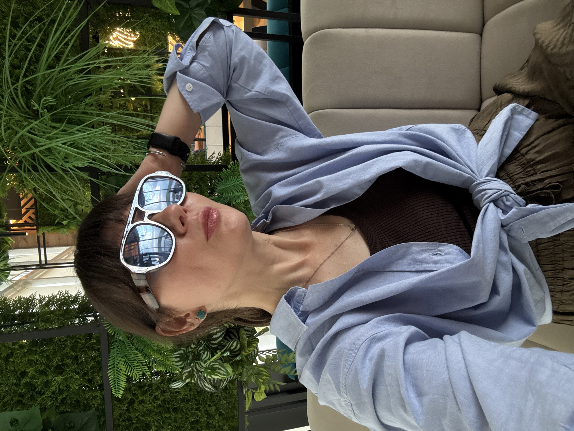 A woman with dark hair and bangs is pictured from the chest up, sitting on a beige sofa. She is wearing a light blue unbuttoned shirt tied at the waist over a brown ribbed tank top, and olive green pants. She has large, silver-rimmed sunglasses with blue lenses on, and is touching the back of her head with her right hand. A silver necklace with a small pendant is visible. She is also wearing square, teal stud earrings and a black watch on her left wrist. The background consists of a wall with artificial greenery and some decorative lighting. The overall scene appears to be indoors, possibly a cafe or a lounge, with a modern and lush aesthetic. The specific location in Chaikovsky, Russia, is not discernible from the image itself.