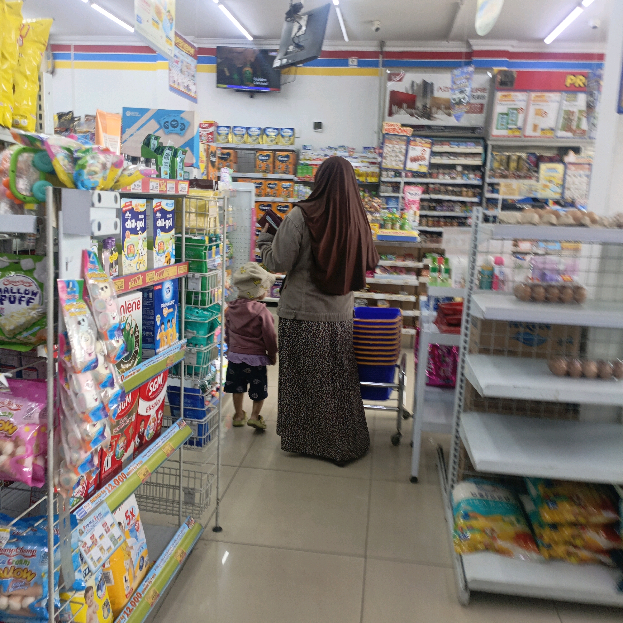In Salatiga, Indonesia, a woman in a brown hijab and a patterned skirt stands in a well-lit convenience store, holding a book. Beside her, a small child wearing a pink jacket, a yellow sunhat, and dark shorts faces away from the camera. The store shelves are stocked with various products, including packaged food items, drinks, and cleaning supplies. A television screen is mounted on the wall above a shelf displaying more products. The floor is tiled.