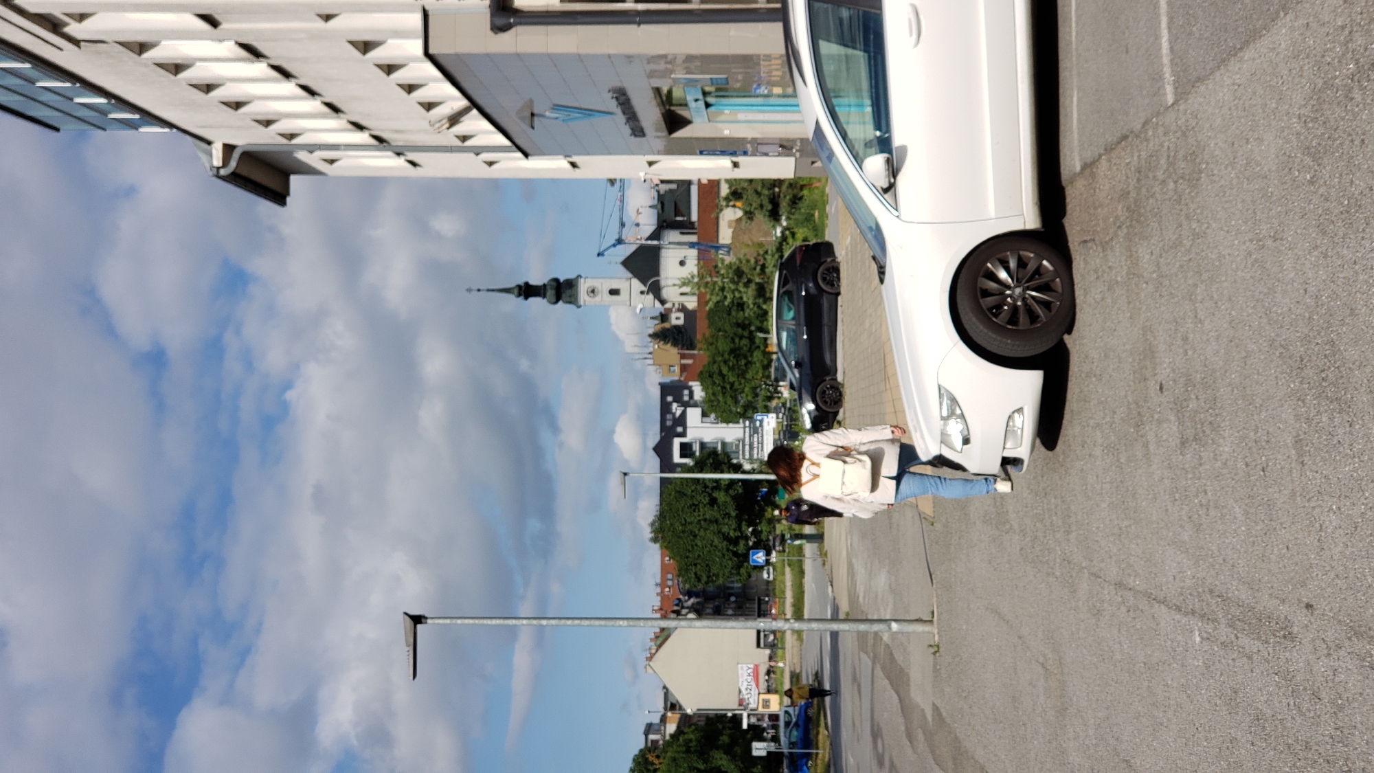 The image captures a street scene in Prešov, Slovakia, likely on a bright, cloudy day. In the foreground, the right side of a white sedan is visible, parked on asphalt. A woman with a light-colored backpack and blue jeans is walking away from the camera, towards the left of the frame. She is walking on the asphalt, near the car. Behind her, a black SUV is parked. Further in the background, a streetlamp stands to the left of the frame, and several buildings are visible, including a modern white building on the right with a blue logo, and a church with a prominent dark steeple in the center-left distance. The sky is filled with scattered white clouds against a blue backdrop. Construction cranes are also visible behind the church. A blue car is parked on the far left.