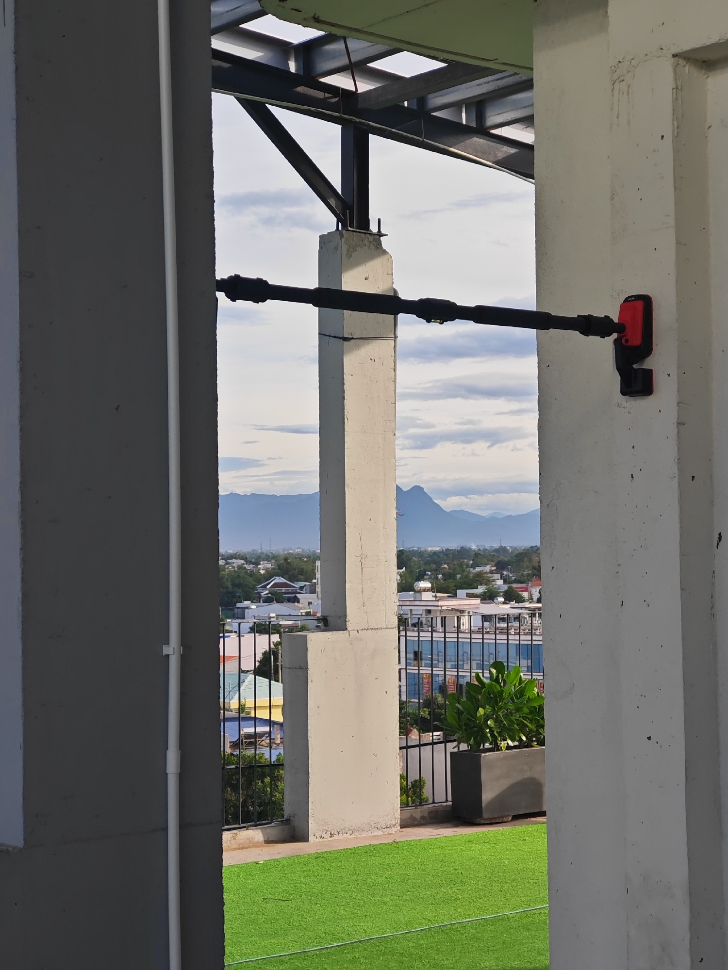 The image captures a view through an opening framed by two concrete pillars. In the foreground, artificial turf covers the ground, appearing bright green. To the right, a large concrete planter holds a lush green plant. Beyond the pillars, a cityscape unfolds with various buildings, some exhibiting colorful facades. A metal fence separates the rooftop area from the buildings below. In the distance, a mountain range is visible against a cloudy sky. A black horizontal pipe, likely part of a structure or utility, stretches across the frame. Attached to the right pillar, a red and black device is mounted on the pipe. The scene appears to be on a rooftop, possibly in a commercial or residential area of Đà Nẵng, Vietnam, given the architectural style and the mountainous landscape in the background, which is characteristic of the region surrounding the city.