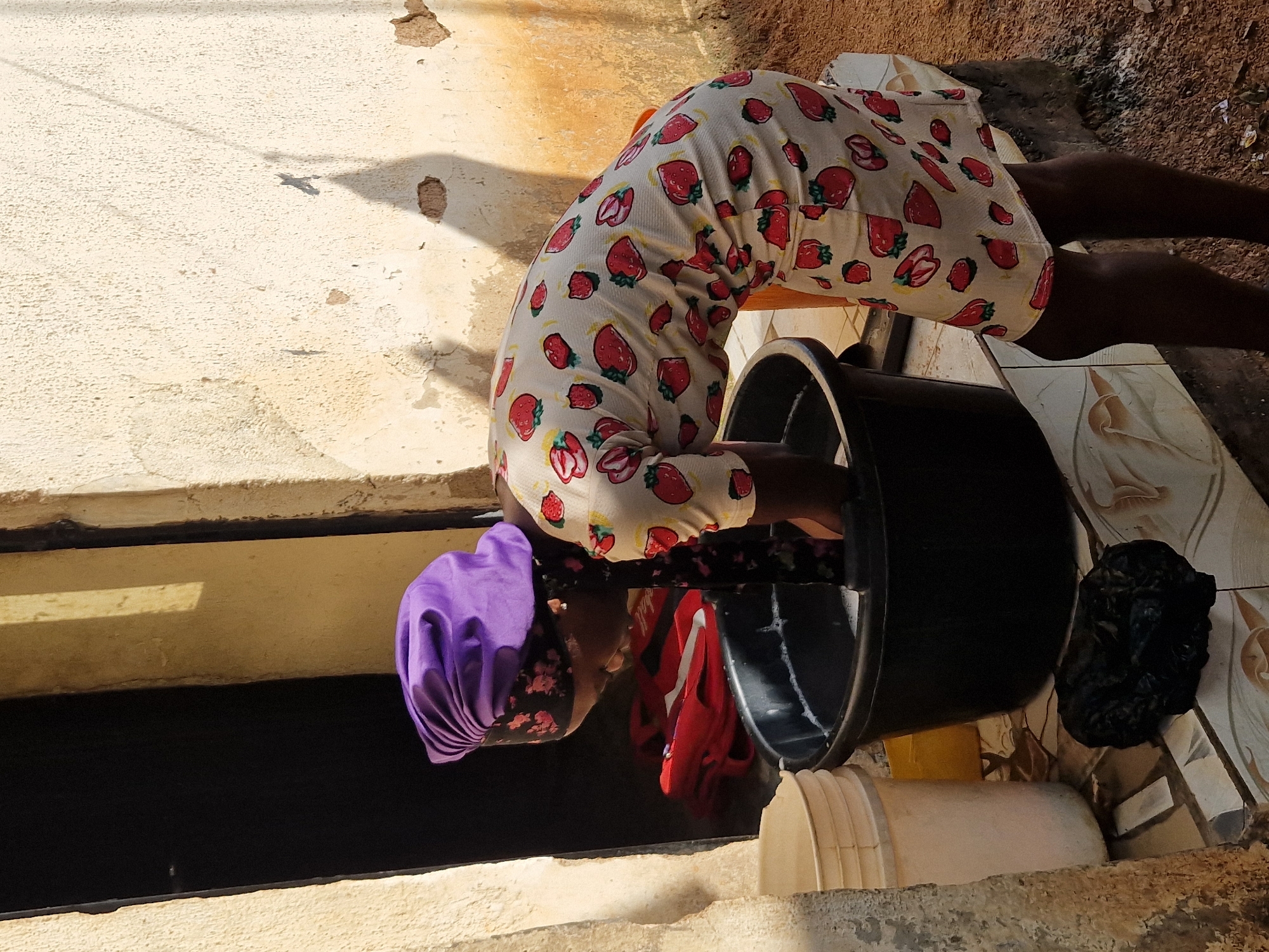 A woman wearing a purple headscarf and a white dress with a strawberry print is washing clothes in a black plastic basin filled with soapy water. She is bent over the basin, with her hands submerged in the water. To her left is a light-colored plastic bucket. Behind her, to the left, is an open doorway revealing darkness, and a stack of folded red clothing. The scene appears to be outdoors, with a rough, textured wall behind the woman, and a tiled surface on the ground. The environment suggests a humble setting, possibly in a residential area of Jos, Nigeria, given the architectural style and outdoor washing activity. Sunlight casts shadows on the wall and ground.