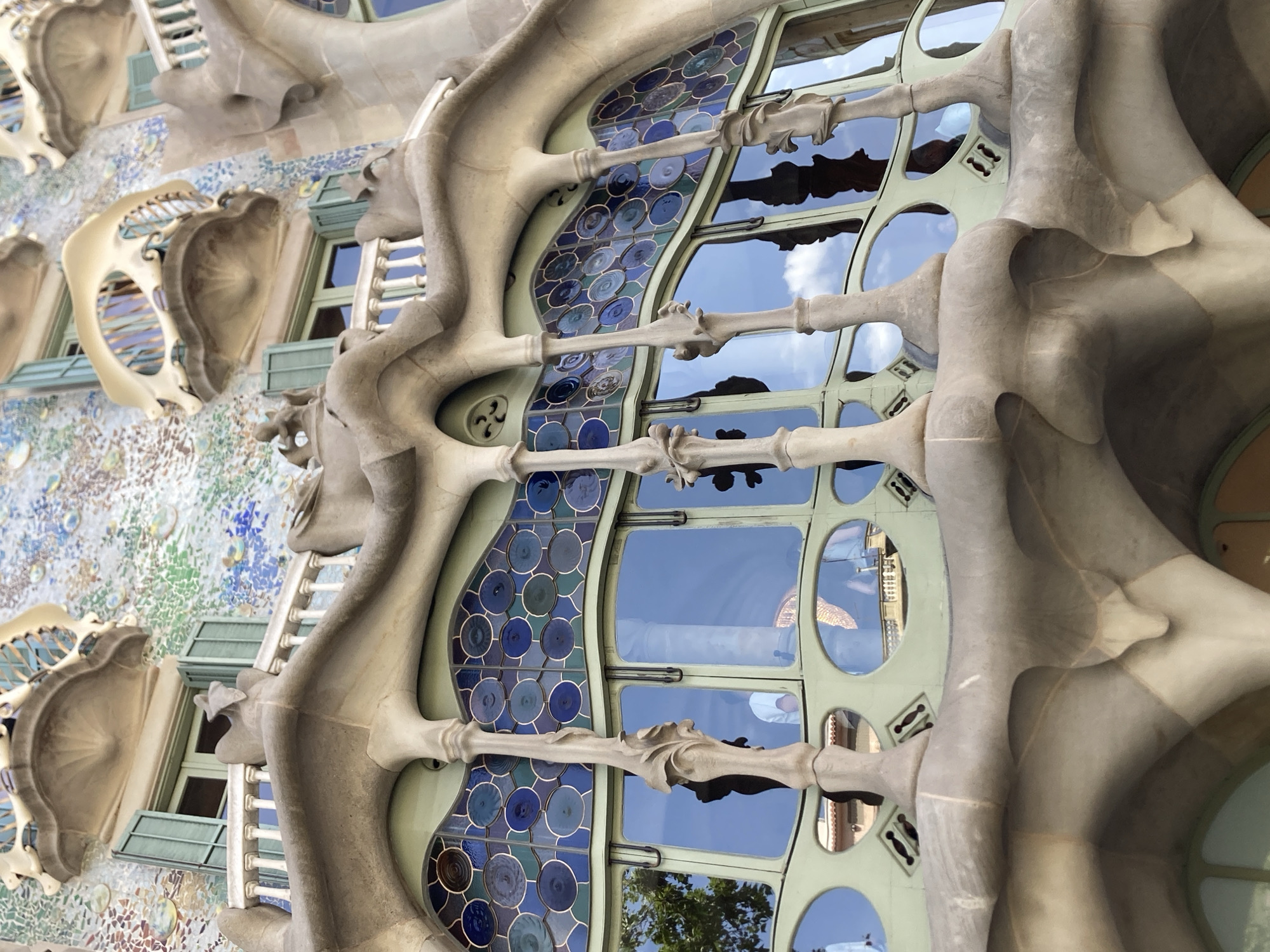 This is a detail shot of the facade of Casa Batlló in Barcelona, Spain, a building designed by Antoni Gaudí. The image focuses on a section of the building's distinctive organic, sculptural elements, with a large window as the central subject. The window is divided into panes framed in a light green, with a decorative band of circular blue mosaic tiles running vertically along its left side. The mosaic tiles vary in shade from deep navy to lighter blues and greens, with some appearing to be iridescent.
The window panes reflect a clear blue sky with scattered white clouds, suggesting a bright, sunny day. The sculptural elements framing the window are a pale, stone-like material, curving and undulating in a way that resembles bone or natural formations. These organic shapes are repeated throughout the facade, with bone-like protrusions extending horizontally across the window, creating a unique architectural aesthetic.
Below the main window, there are glimpses of other architectural details, including what appear to be balconies or alcoves with arched openings, also rendered in the same flowing, organic style. The background on the left shows a textured wall covered in a mosaic of various small, colorful tiles in shades of blue, green, white, and pink, contributing to the building's overall whimsical and fantastical appearance. The overall impression is one of artistic innovation and a playful interpretation of natural forms. There is no visible text in the image.