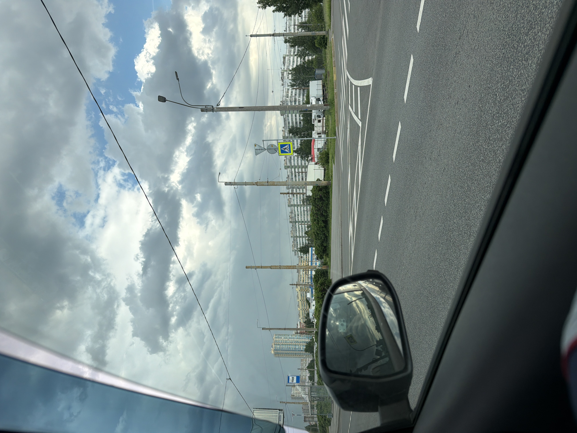 This image presents a daytime view from inside a vehicle, looking out the side window. The sky is largely overcast with large, grey clouds, interspersed with clear patches of blue sky, particularly in the upper left. Overhead, several thin black utility lines traverse the frame.
The foreground features a dark asphalt road marked with distinct white dashed lines and a curving solid white line, indicating road divisions or turn instructions. A portion of the car's side mirror is visible in the lower right, reflecting the cloudy sky and distant elements.
Along the side of the road, several tall utility poles, equipped with streetlights, punctuate the scene. Attached to one of these poles is a prominent yellow and blue pedestrian crossing sign, depicting a white walking figure within a blue triangle. A smaller, rectangular white sign is positioned beneath it, but its details are too indistinct to read.
Beyond the road and poles, green trees line the view, partially concealing numerous multi-story residential buildings, which extend across the horizon. These buildings bear the architectural style common to post-Soviet urban environments. White trucks or trailers are parked near some of these structures in the middle ground. Further in the distance, more slender, tall structures, possibly industrial chimneys or ventilation shafts, pierce the skyline. A small blue and white sign, possibly for a bus stop or an informational point, is also visible but illegible.
The scene depicts an urban or suburban landscape during daytime, with overcast but bright weather. The architectural style and road signage are consistent with a location in Saint Petersburg, Russia. No people are visible in the frame.