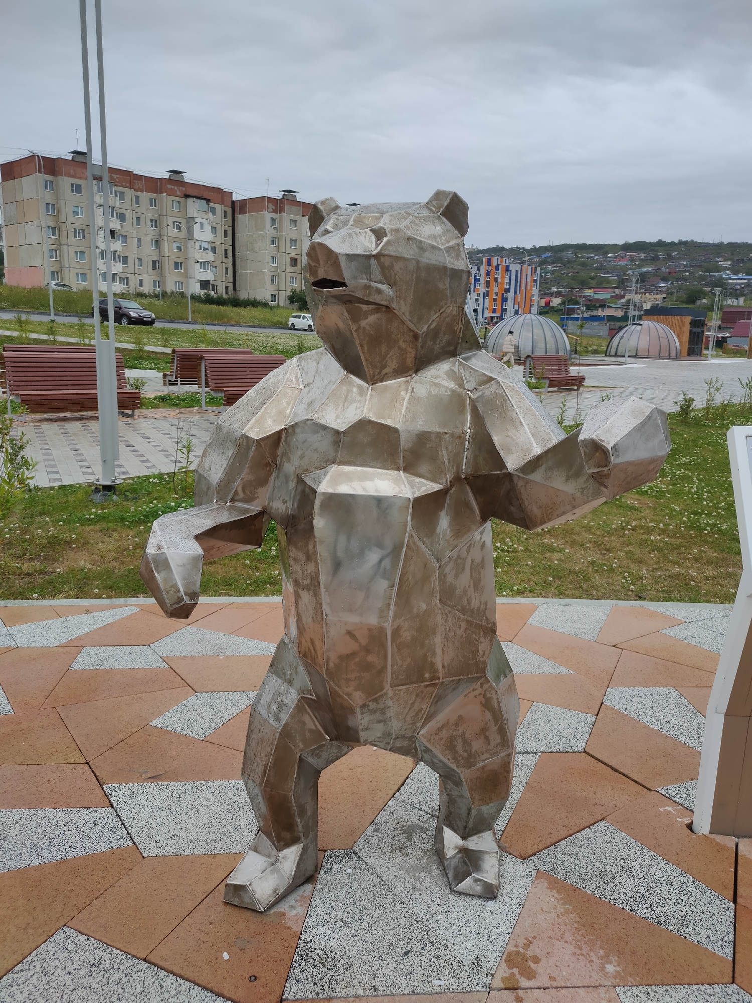 A polished metal sculpture of a bear stands on a patterned paved area. The bear sculpture is polygonal and appears to be made of many flat metal panels. The bear is in a standing position, with its arms extended slightly outward. In the background, there are multi-story apartment buildings and a hilly landscape with scattered structures. The sky is overcast. The paved area consists of reddish-brown tiles and lighter speckled tiles arranged in a geometric pattern. There are benches and greenery around the sculpture. The location is identified as the "City of Magadan, Russia".