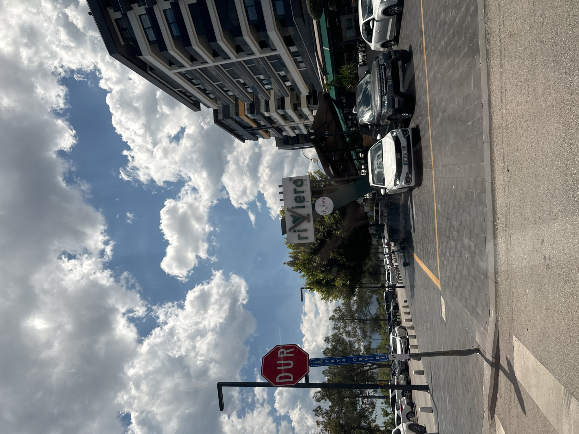 The image shows a street scene in Konyaaltı, Turkey. A building with a sign that reads "Riviera Bistro" is visible. A red stop sign with "DUR" written on it is also present. Cars are parked along the side of the street. The sky is blue with puffy white clouds.