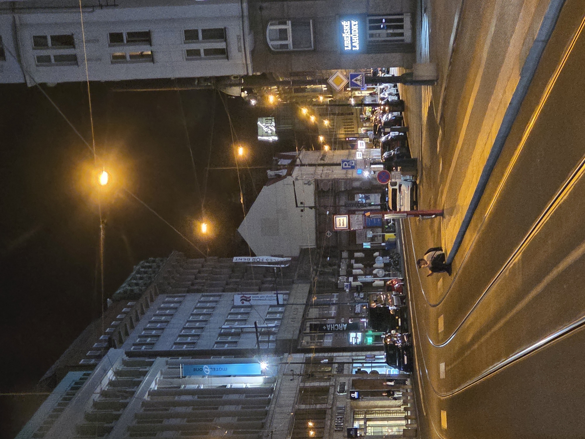 The image depicts a nighttime street scene in an urban environment, viewed from an elevated perspective looking down a main thoroughfare. The sky is dark, and the scene is illuminated by numerous streetlights and glowing signs from businesses.
In the foreground and running diagonally through the image are tram tracks, composed of two parallel sets of rails embedded in the paved road. The road surface appears well-maintained. On the right side of the road, a pedestrian is visible walking towards the left of the frame. To the left of the tram tracks, there's a sidewalk area with parked cars and what appear to be scooters or motorcycles.
Along both sides of the street, multi-story buildings line the thoroughfare. Many windows are visible, some lit from within, suggesting ongoing activity. Various commercial signs adorn the building facades. Notable signs include "LIBEŠSKÉ LAHŮDKY" on an upper-level building, "MOTEL ONE" on a bright blue sign on a modern-looking building, and "ARCHA+" on another sign. Further down the street, a sign reads "OFFICES FOR RENT". Another partially visible sign reads "FLORENTINUM".
Standard European traffic signs are scattered throughout the scene, including a yellow diamond indicating a priority road, a blue square with a white 'P' for parking, and a red circular sign, likely indicating a prohibition or restriction. The overall impression is that of a bustling city street after dark.