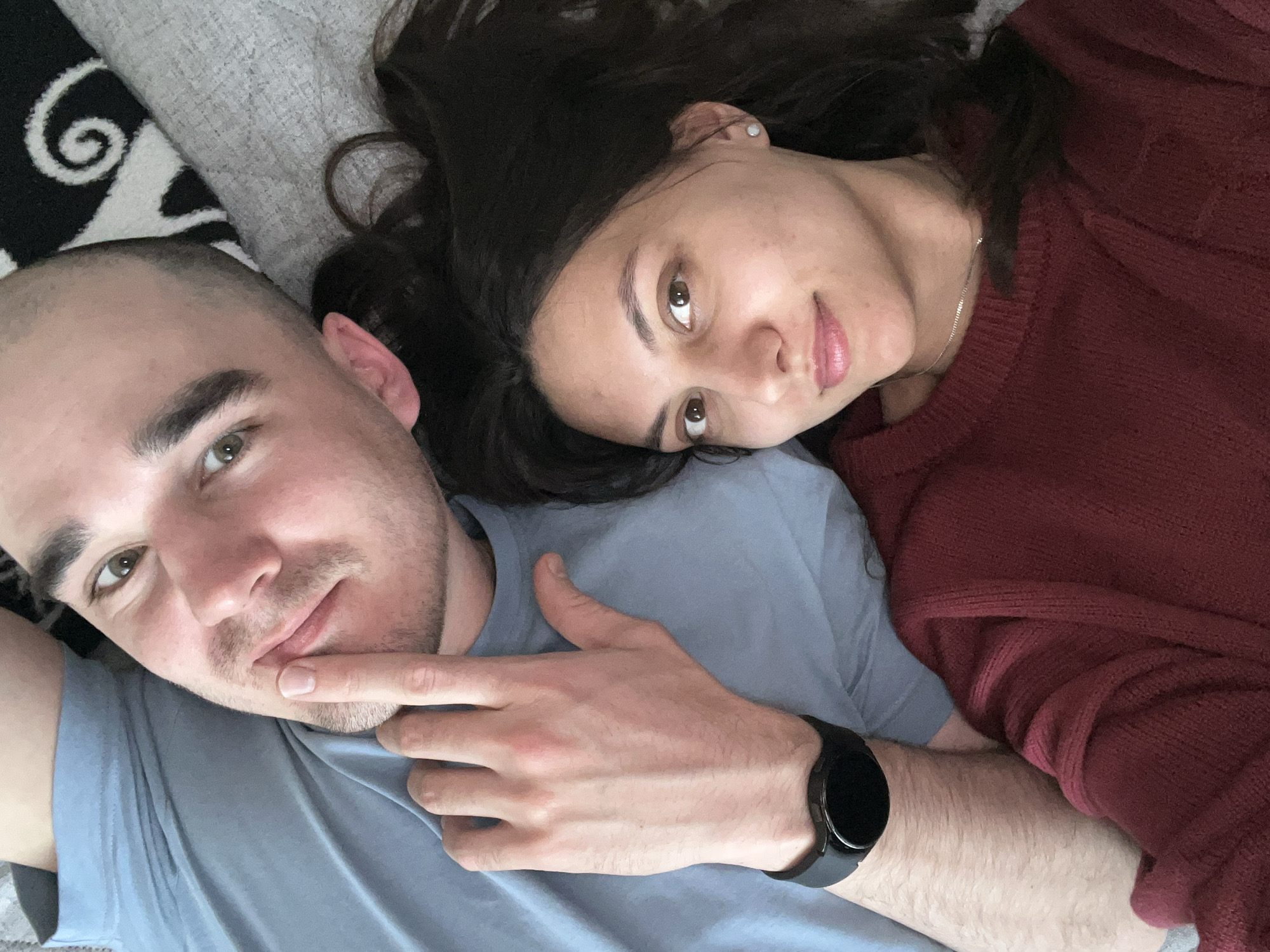 A selfie-style image captures two individuals lying close together on what appears to be a bed or couch. The lighting is natural, and the focus is sharp on the faces of the two individuals.
The first individual, positioned closer to the camera, is a male with a shaved head and light skin. He has light-colored eyes and is wearing a light blue t-shirt. His right hand is raised, with his index finger lightly touching his lips. A black smartwatch is visible on his left wrist.
The second individual, a female, is lying with her head resting on the male's chest. She has long, dark hair and dark eyes. She is wearing a burgundy sweater and a delicate silver necklace. A small stud earring is visible in her ear.
The background includes a textured gray surface and a black and white patterned cushion.
There is no indication of the location being in the city of Yekaterinburg, Russia.