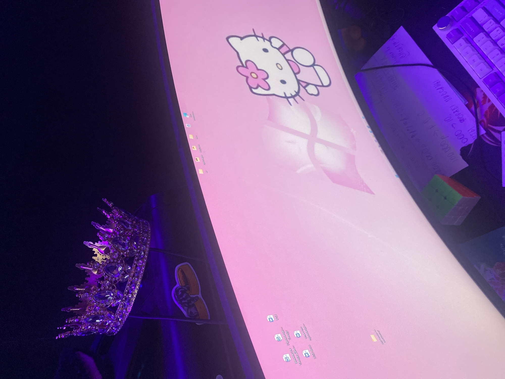 The image captures a dimly lit computer workstation, likely at night, imbued with a strong purple ambient light. A large, curved monitor displays a cheerful pink wallpaper featuring a prominent Hello Kitty graphic. The monitor's desktop shows numerous file icons, many appearing to be Microsoft Word documents, with titles written in Russian, such as "Машинку," "Бикмасов Производст," "Тимур Дне," and "PA3 docx."
To the right of the monitor, a white mechanical keyboard is visible. Adjacent to it, a piece of paper with handwritten notes is present, illuminated by the purple light. The legible text on the paper includes Russian phrases like "ЧТЫ ФОРАБЫ ПИССЛИ" (possibly "чтобы форумы писали" - for forums to write), numerical sequences like "000-10," and "100.000 POC" (likely indicating Russian rubles). A multi-colored Rubik's Cube rests near the notes.
On the left side of the frame, a glittering, jeweled crown, primarily purple and gold, is prominently displayed. Below it, a playful sticker depicting a cat in a hot dog bun is affixed to a dark surface. The setup conveys a personalized and cozy environment, with the Russian text reinforcing the context of Izhevsk, Russia.