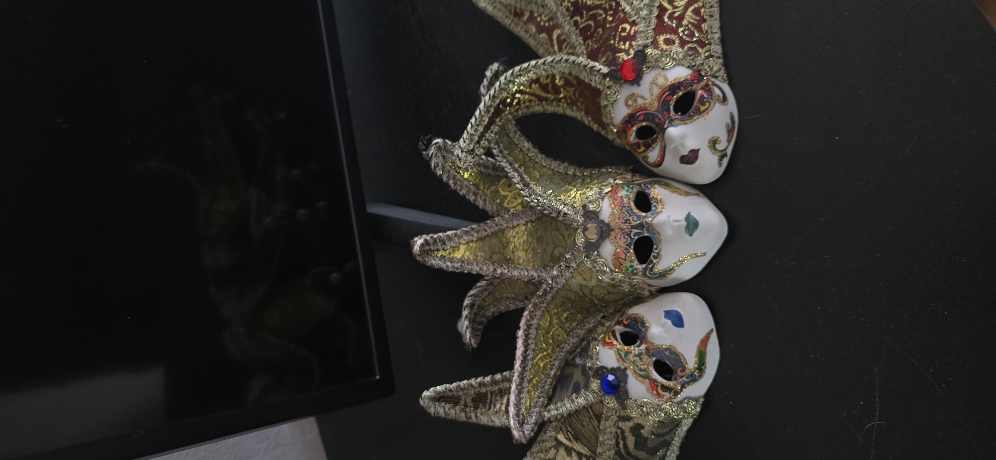 The image depicts three Venetian masks lying on a dark, possibly black, surface. The masks are arranged vertically, one atop the other, with a portion of a black-framed object (possibly a monitor or TV) visible on the left. The masks are ornate, with white faces and decorative elements in gold, red, and various other colors. They feature designs around the eye holes and other facial decorations, with each mask having a unique overall design. The masks appear to be in a stacked or layered arrangement. The precise location in Varna, Bulgaria is not determinable from the image alone.