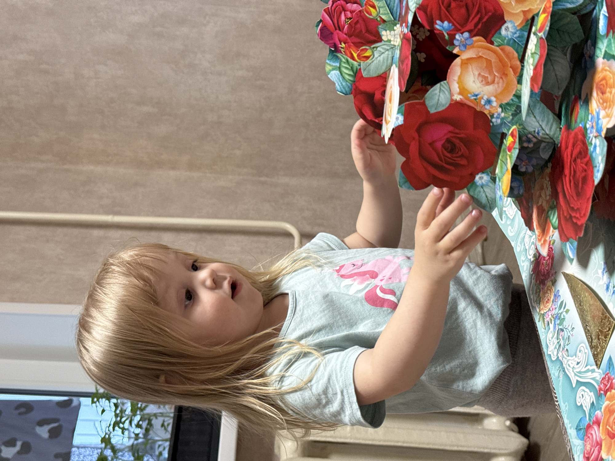 A young child with blonde hair, wearing a light blue shirt with a pink unicorn design and gray pants, is touching a large paper flower arrangement. The arrangement is filled with red, orange, and blue flowers. The child is positioned near a window with visible greenery outside. A radiator is visible in the background. The wall behind the child is beige. The location is indoors, possibly a home. There is no other information on location.